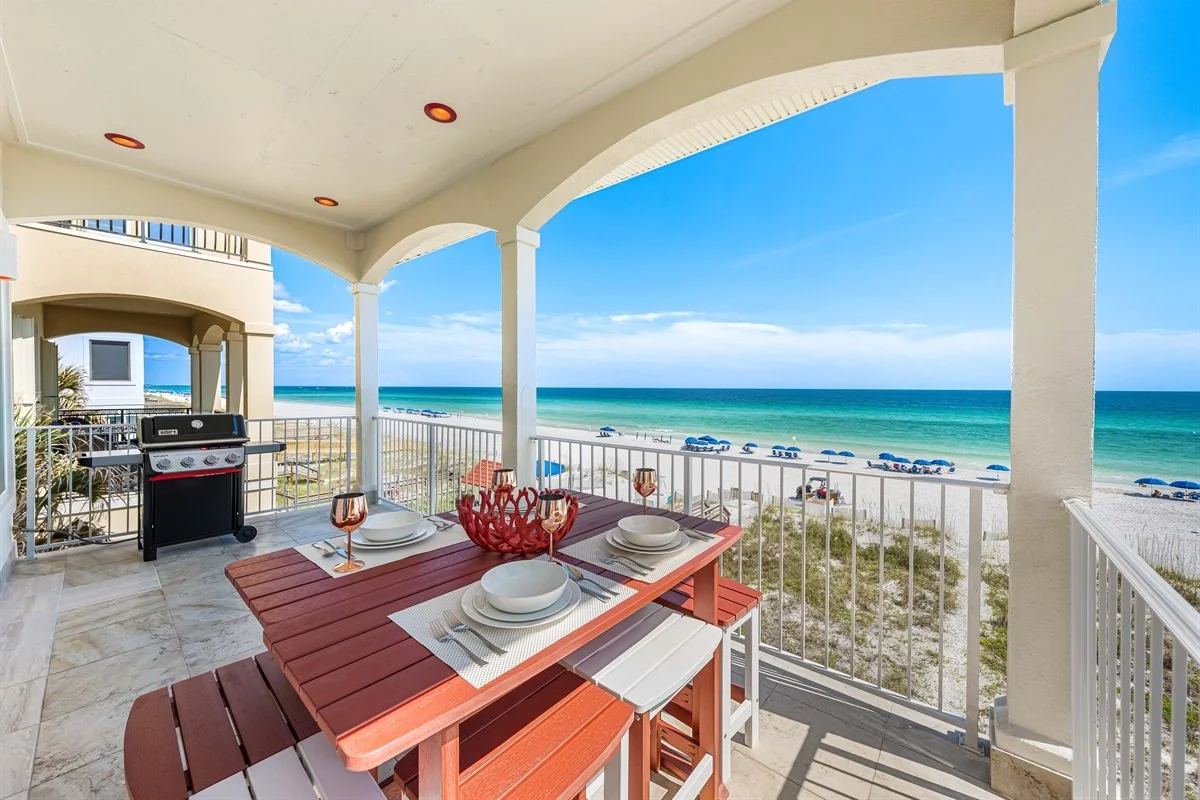 Beachfront balcony with a dining table, set for four, overlooking the ocean, with lounge chairs on the sandy beach and umbrellas in the distance