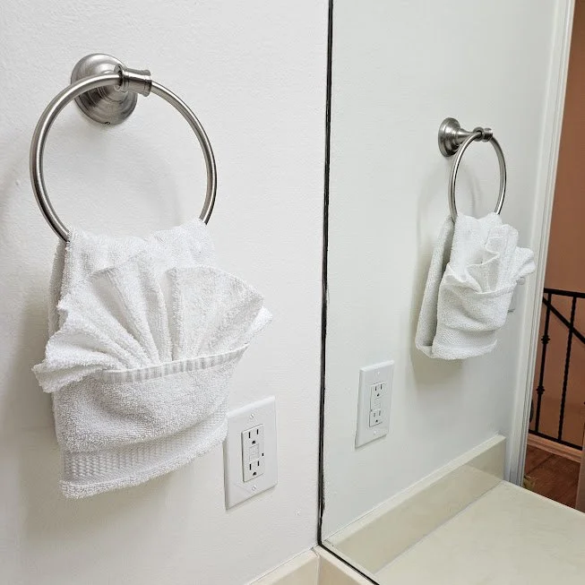 A white hand towel folded into a fan shape hanging on a silver towel ring next to a mirror. There is an electrical outlet below the towel ring.