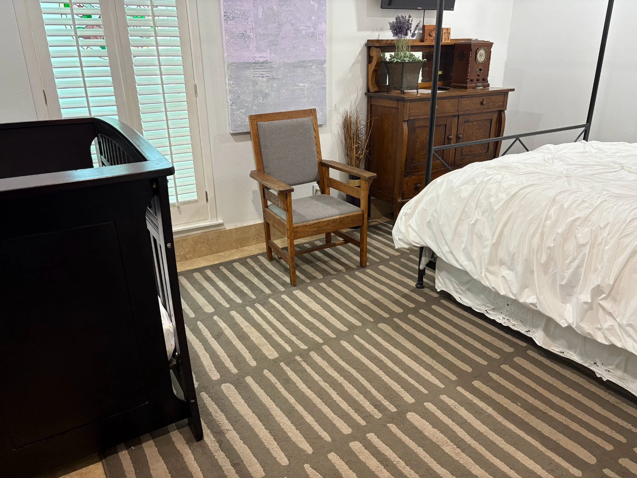 A bedroom corner with a bed, a wooden chair, a wooden cabinet with decorative items, a potted plant, and a striped rug, with a wall-mounted TV and window blinds in the background.