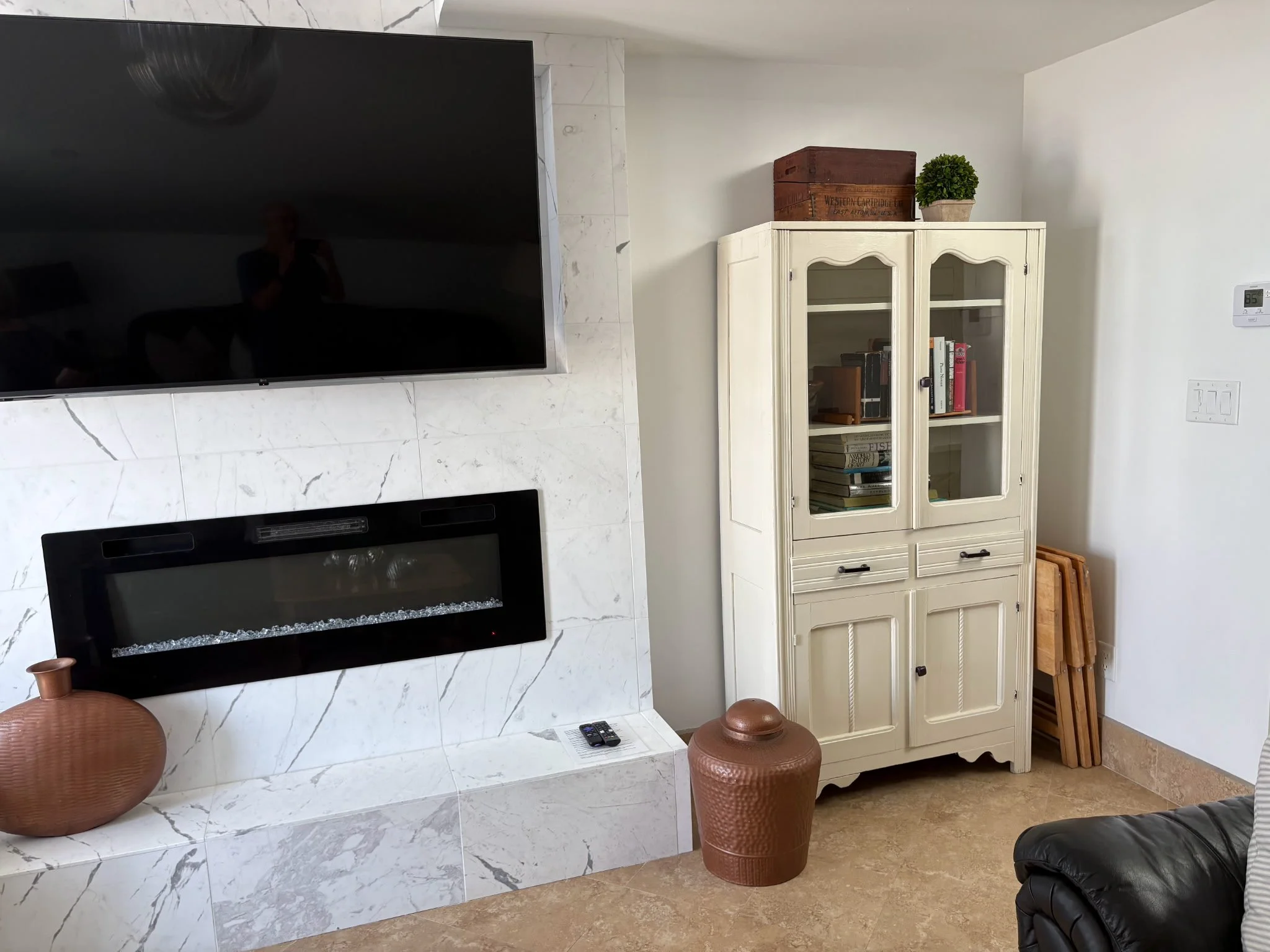Living room corner with a flat-screen TV mounted on a white marble-effect wall, a fireplace below, and decorative items including a large terracotta pot and a textured brown jar. To the right, a cream-colored cabinet with books and a potted plant on top. A digital thermostat is on the wall, and part of a black leather sofa is visible on the right.