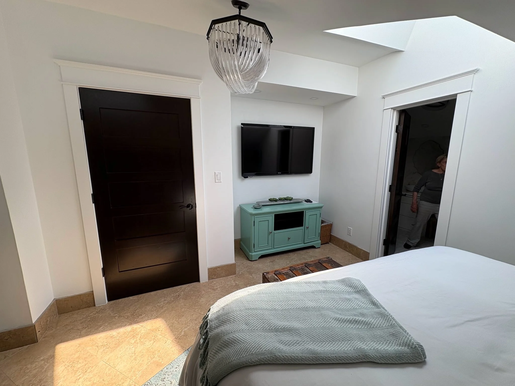 A bedroom with a bed, a TV mounted on a white wall, a black door, and a woman in a grey shirt standing in a doorway. There is a light green cabinet under the TV and a wicker basket on the floor.