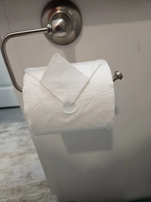 A toilet paper roll on a toilet paper holder with one sheet pulled out and folded over, with a small square paper under the folded sheet.