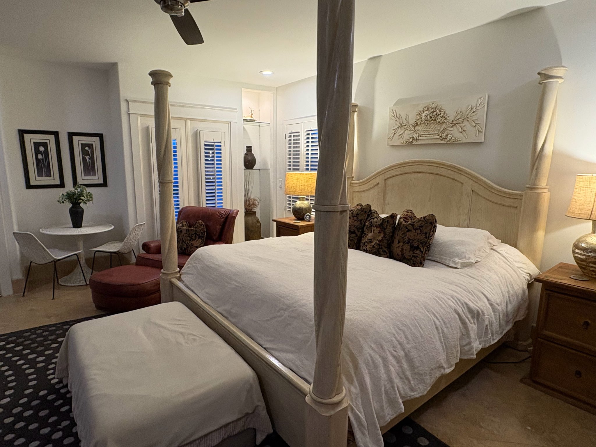 A bedroom with a four-poster bed with white bedding, a wooden nightstand with a lamp, and decorative pillows. There are two framed black-and-white floral pictures on the wall, a small round table with a plant, a chair, and tall windows with white shutters.