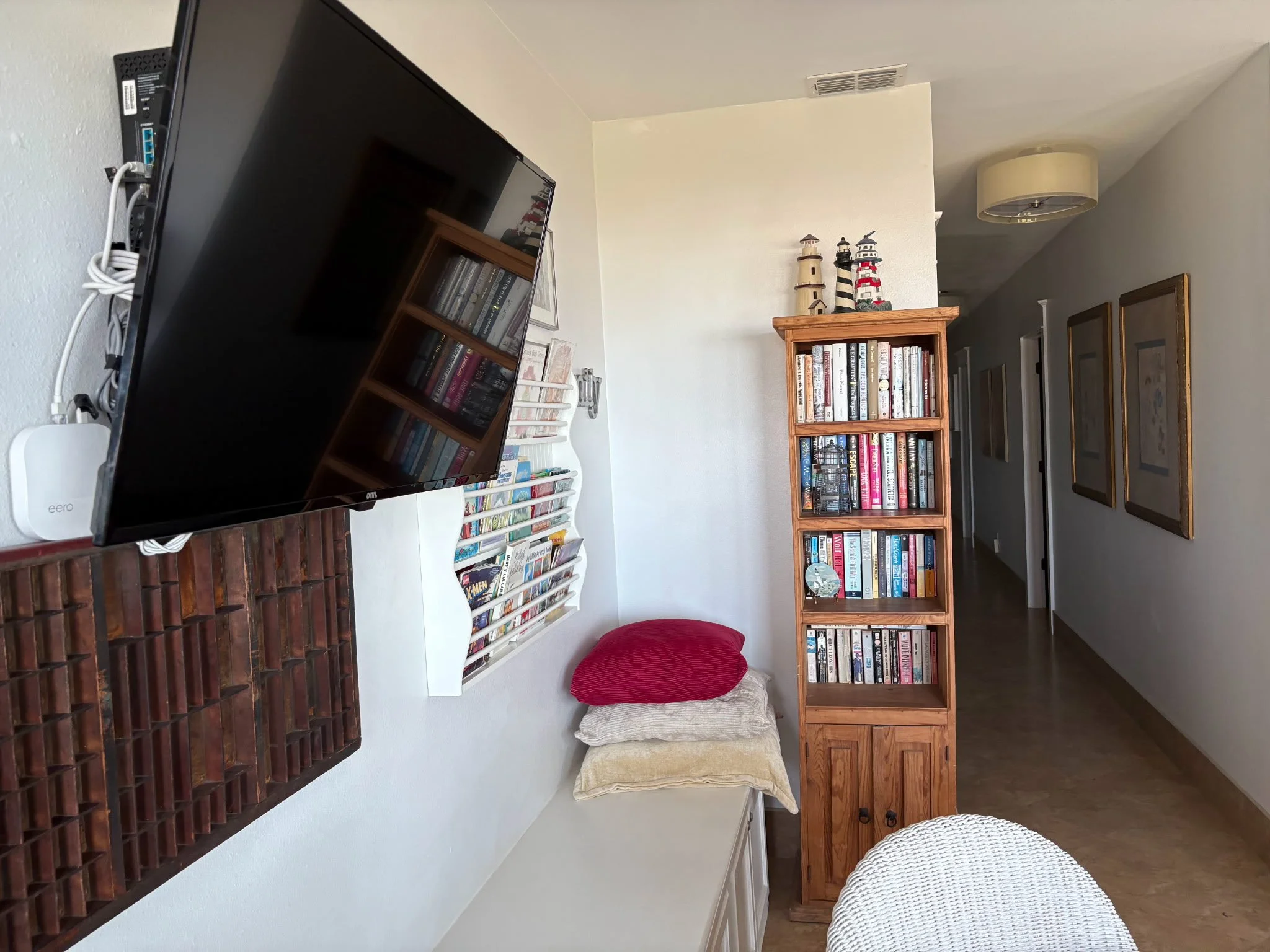 Living room with a flat-screen TV on a wooden stand, a white decorative bookshelf filled with books, a red cushion, a beige cushion, and framed artwork on the wall.