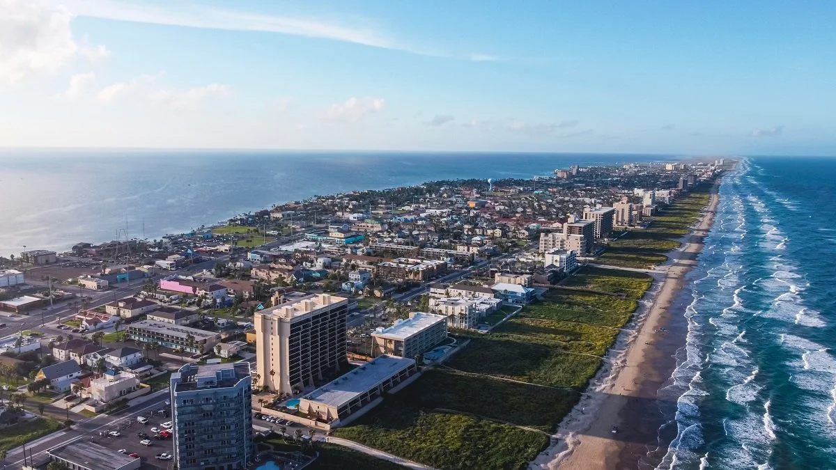 Aerial view of a coastal city with residential and commercial buildings along the shoreline, waves hitting the beach, and a clear sky.