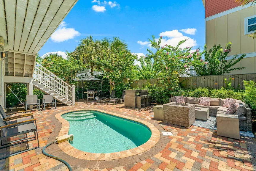Backyard patio with a small kidney-shaped swimming pool, outdoor seating area with cushioned chairs and a sofa, trees, flowers, and a barbecue grill, under a blue sky with some clouds.