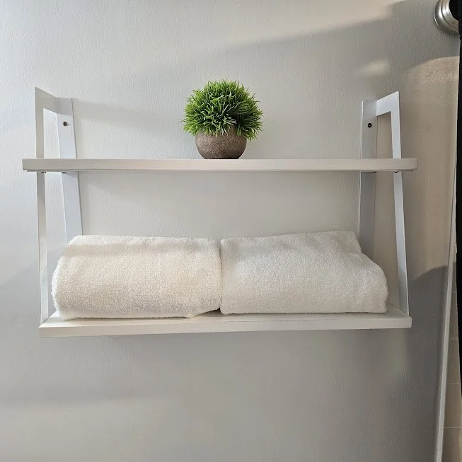 White wall-mounted shelf with a small green plant in a gray pot on top and two folded white towels on the bottom shelf.
