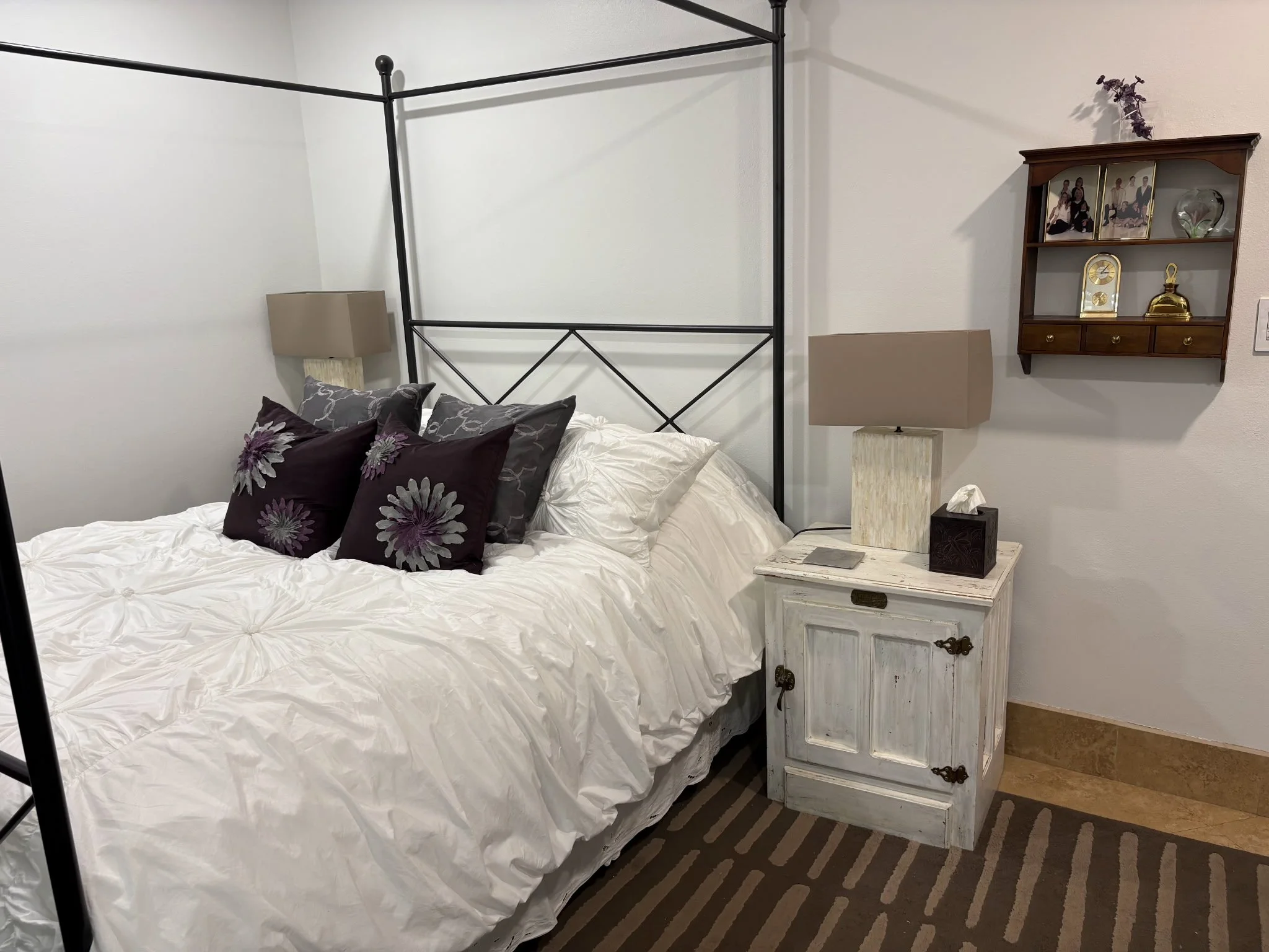 A bedroom with a bed covered in white bedding, decorated with purple and gray floral pillows. There are two beige lamps on either side of the bed, a white distressed nightstand with a tissue box and a small book. On the wall, there is a wooden shelf with family photographs, a clock, a vase, and decorative items.