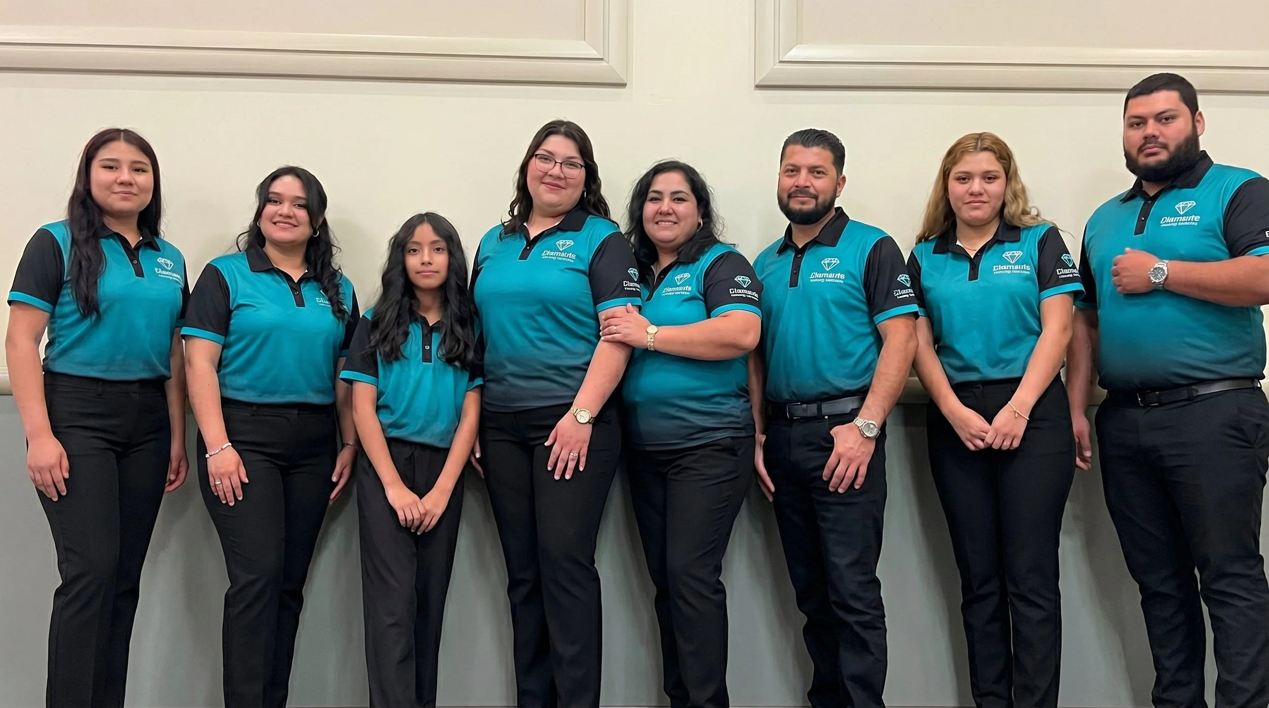 Group of eight people wearing teal and black uniforms standing against a light-colored wall.