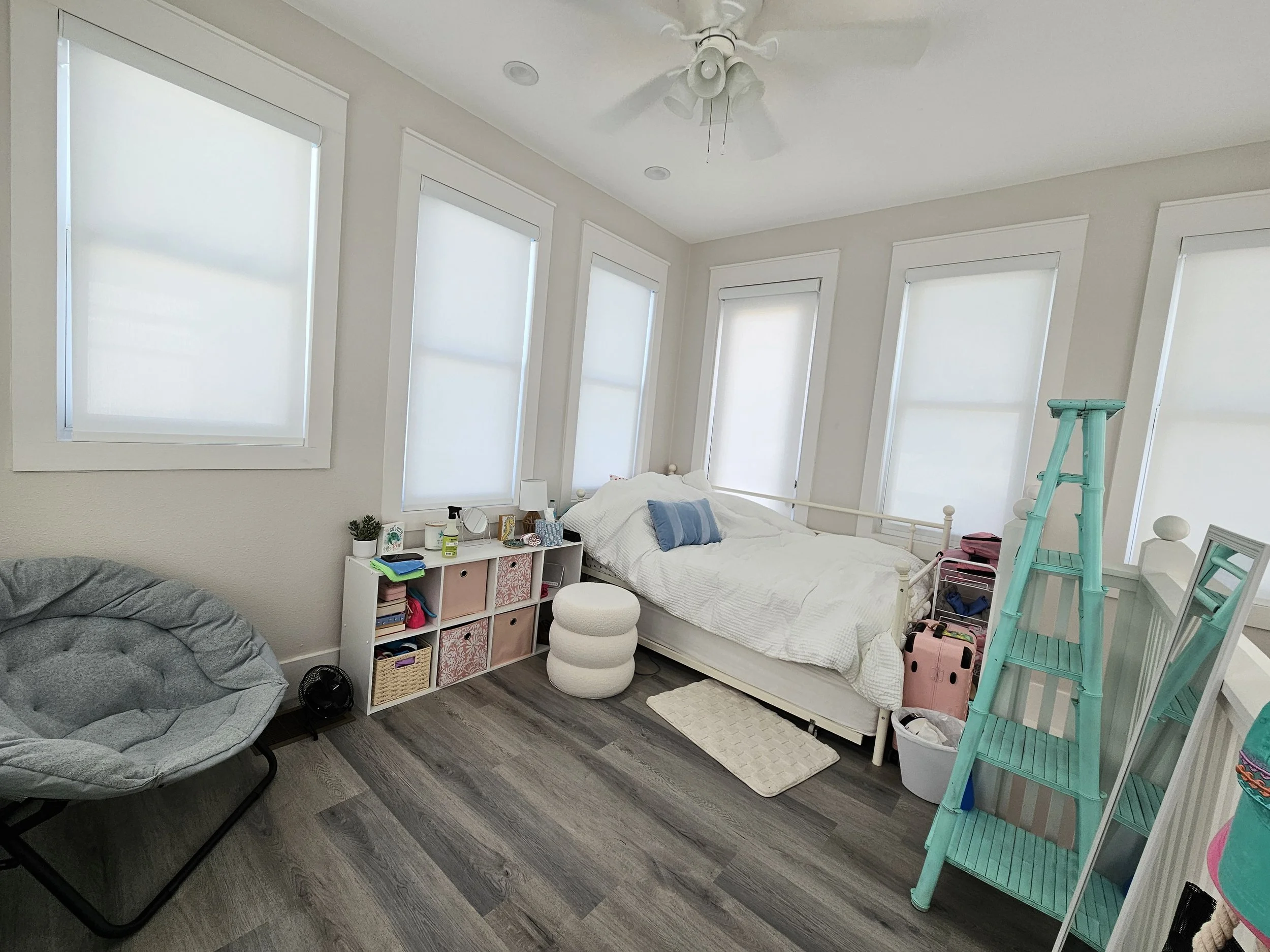A bedroom with six large windows with white blinds, a white bed with a blue pillow, a white cubby shelf with various items, a gray papasan chair, a white ottoman, a teal step ladder, a small pink suitcase, and a white basket, all on dark wood flooring.