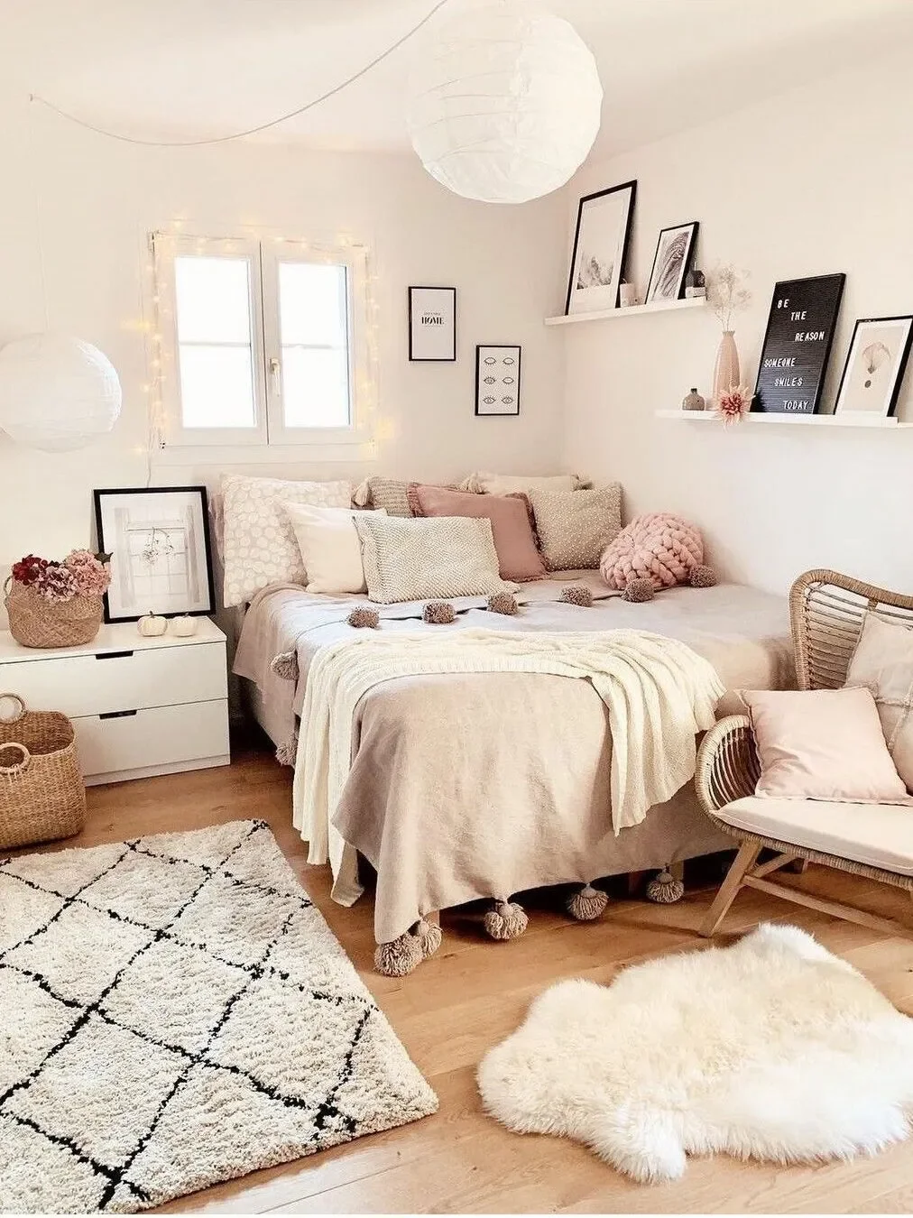 A cozy, neutrally decorated bedroom with a bed against the wall, adorned with various pillows and a throw blanket. There are two fluffy rugs on a wooden floor, a wicker chair with a cushion, and decorative wall art on white walls. The room is softly lit, featuring hanging paper lanterns and string lights around a window, with shelves displaying framed pictures and decor items.
