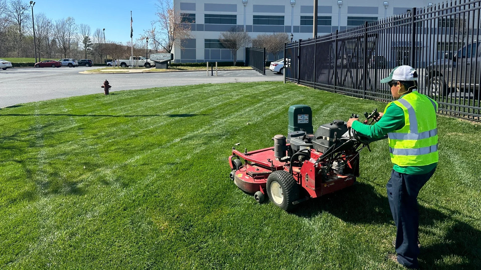 A person operating a ride-on lawn mower cutting bright green grass on a lawn near a black metal fence and a parking lot with parked cars and a large white building in the background.