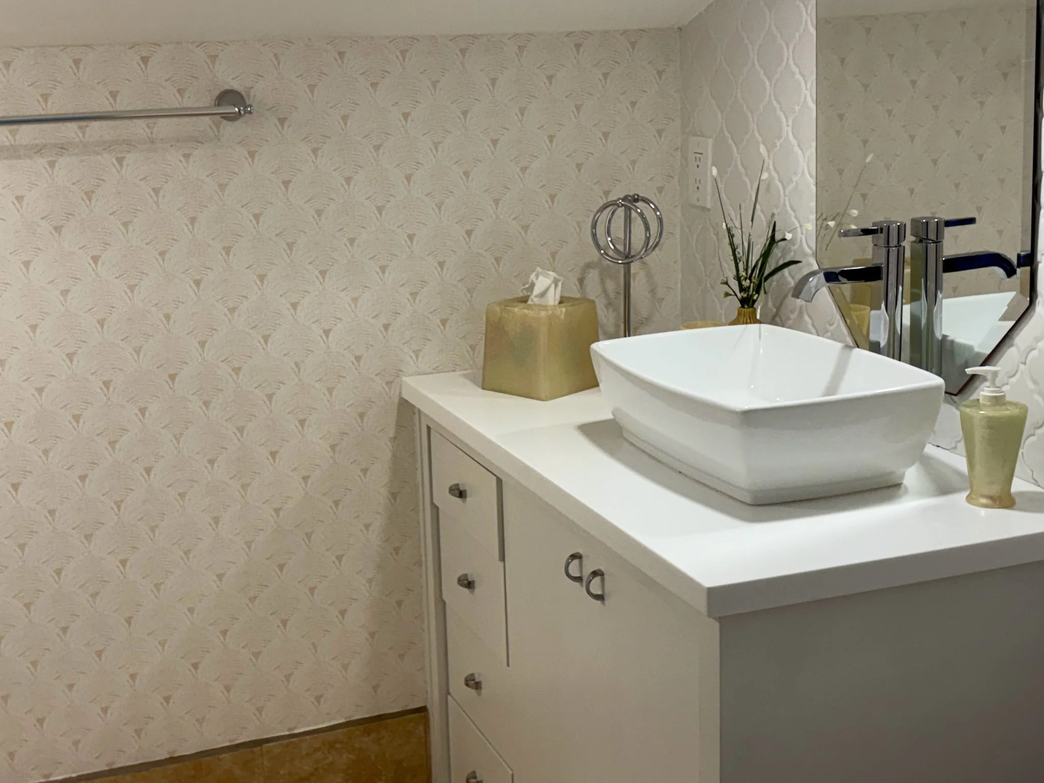 A bathroom with cream-colored textured wallpaper, a white vanity with a vessel sink, a large mirror, two chrome faucets, a small potted plant, a tissue box, a soap dispenser, and decorative metal rings on a stand.