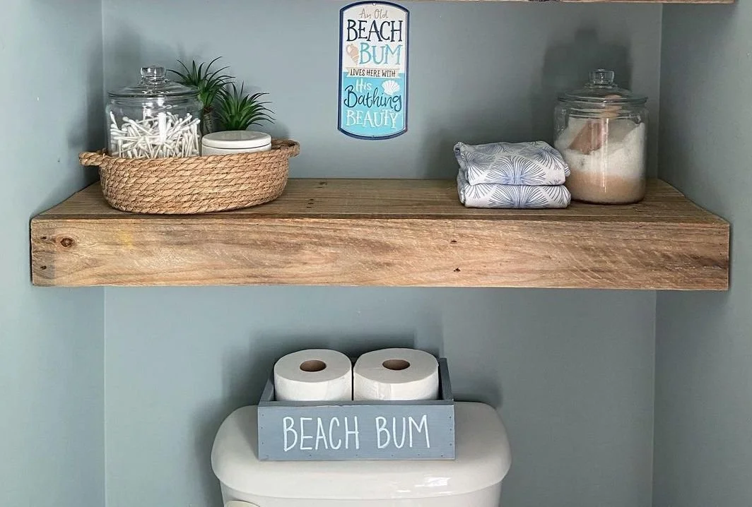Decorative bathroom shelf with jars, towels, a plant, a sign, and a holder with toilet paper rolls that says "Beach Bum".