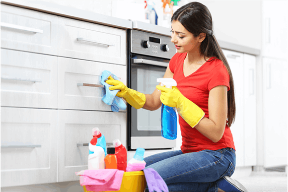 Young woman cleaning kitchen cabinets with spray bottle and cloth, wearing yellow gloves, surrounded by cleaning supplies.