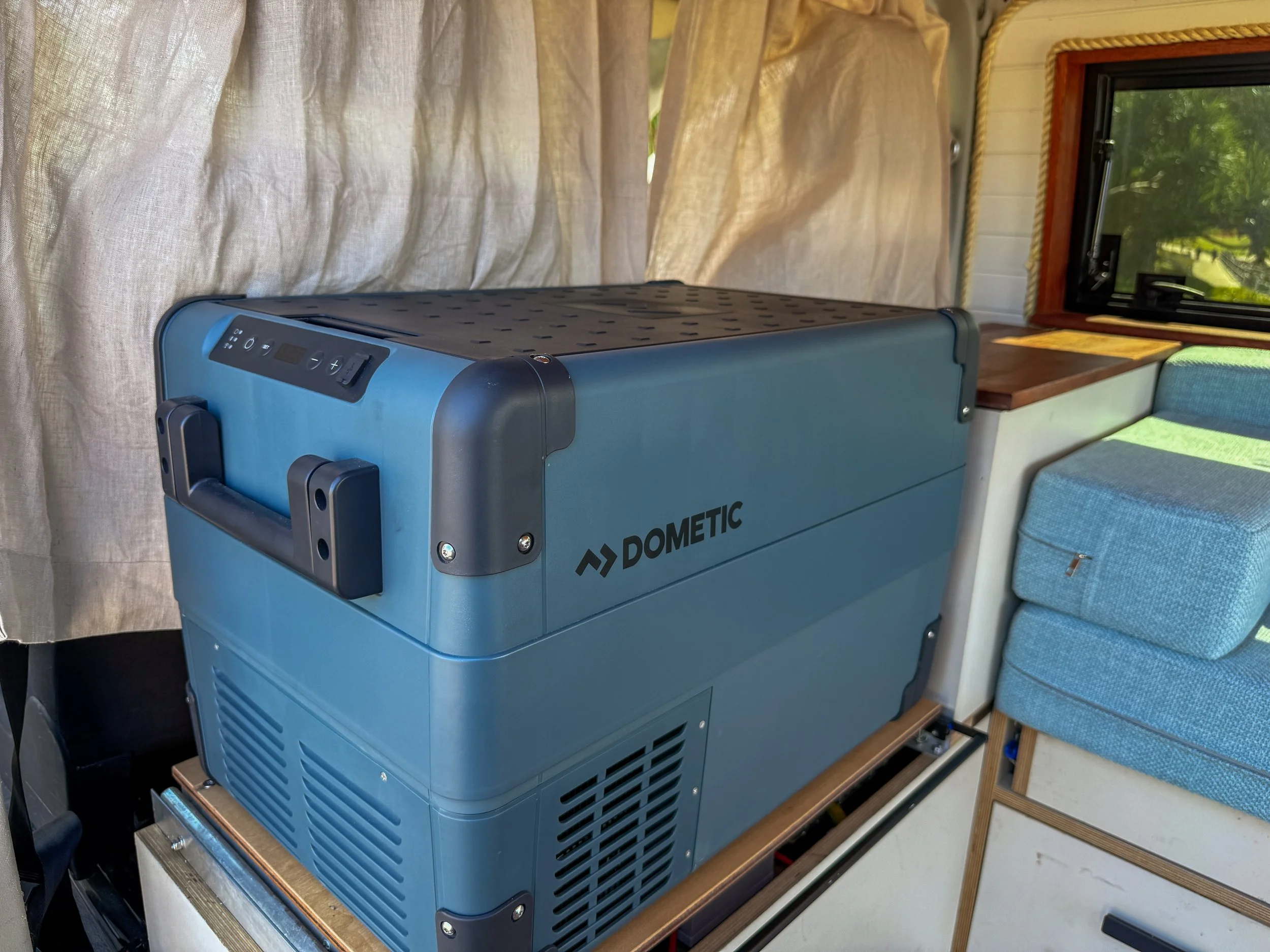 Blue portable refrigerator or cooler with control panel situated inside a cozy room near a window with curtains and a seating area.