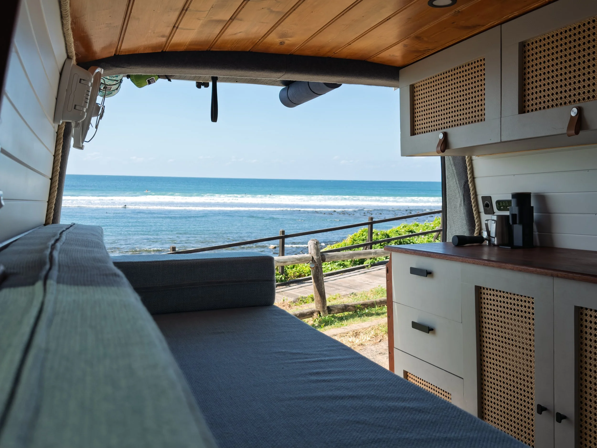Inside a cozy camper van with a view to the beach and ocean through the open back doors. The interior features wooden panels on the ceiling, a gray sofa with striped pillows, a small countertop with a black coffee maker, and a dresser with cane-front drawers. Outside, there's a deck with a wooden railing and lush green plants.