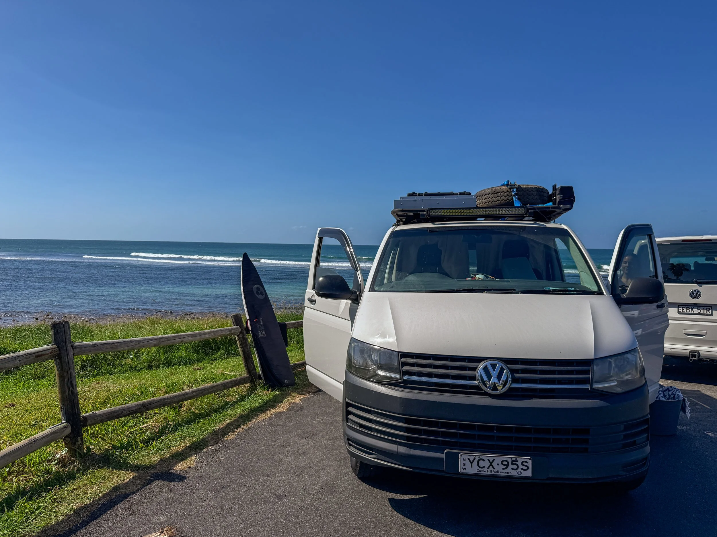 A white Volkswagen van parked near a coastline with ocean waves and a clear blue sky, a surfboard leaning against a wooden fence nearby, and a beach environment.
