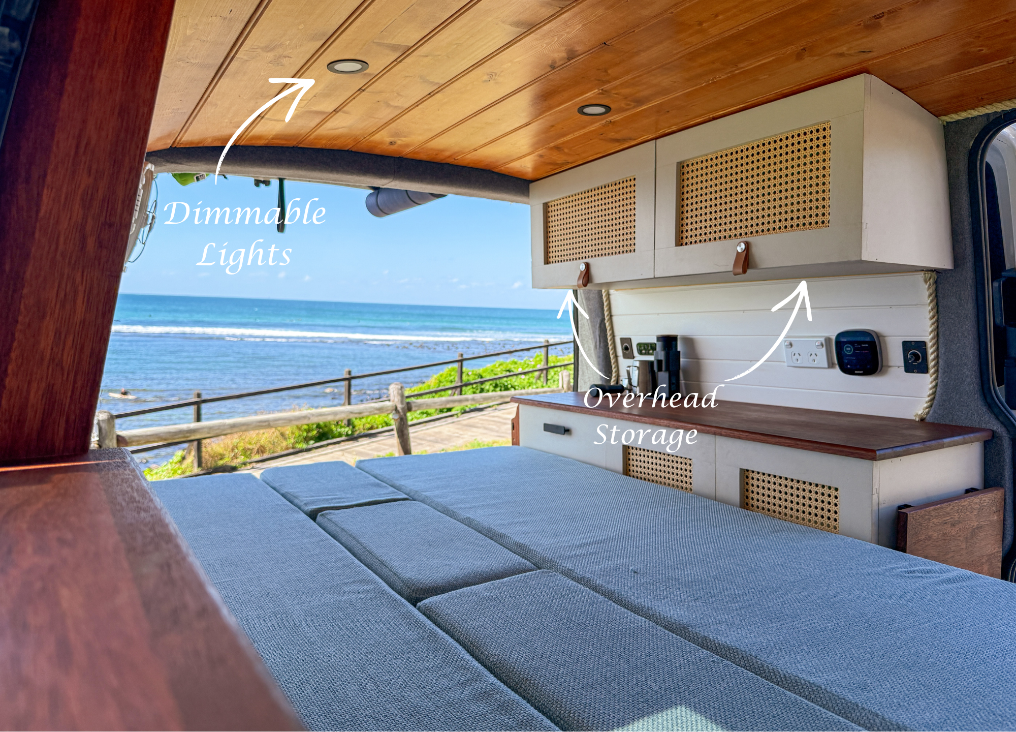 Interior view of a camper van with a view of the ocean, showing a wooden ceiling with recessed dimmable lights, overhead storage cabinets with cane paneling, and a seating area with blue cushions.