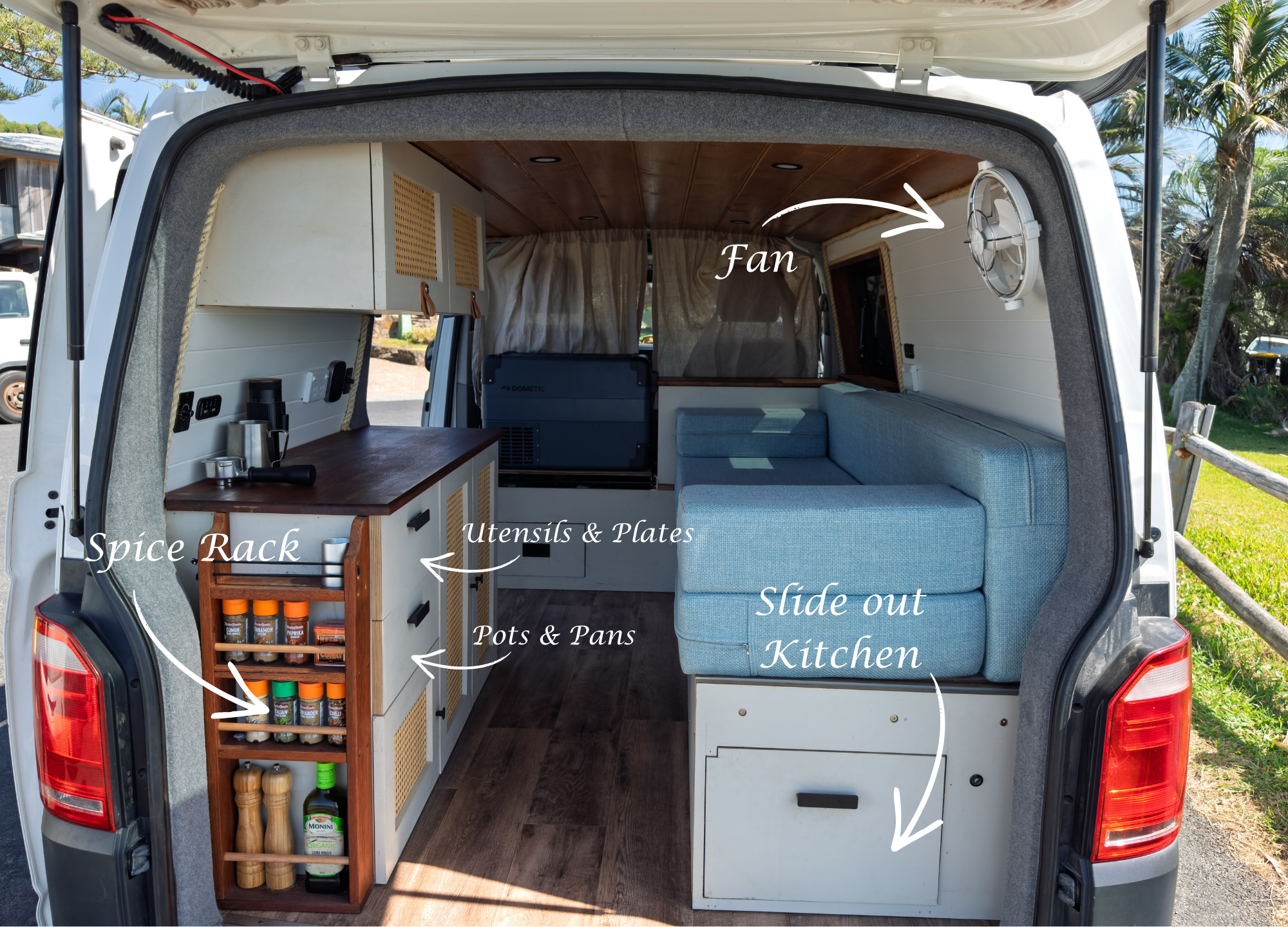The interior of a camper van converted into a small kitchen with a slide-out kitchen area, including a spice rack, utensils and plates, pots and pans, a fan on the right wall, and a seating area with a cushion. Wooden ceiling and flooring, with exter