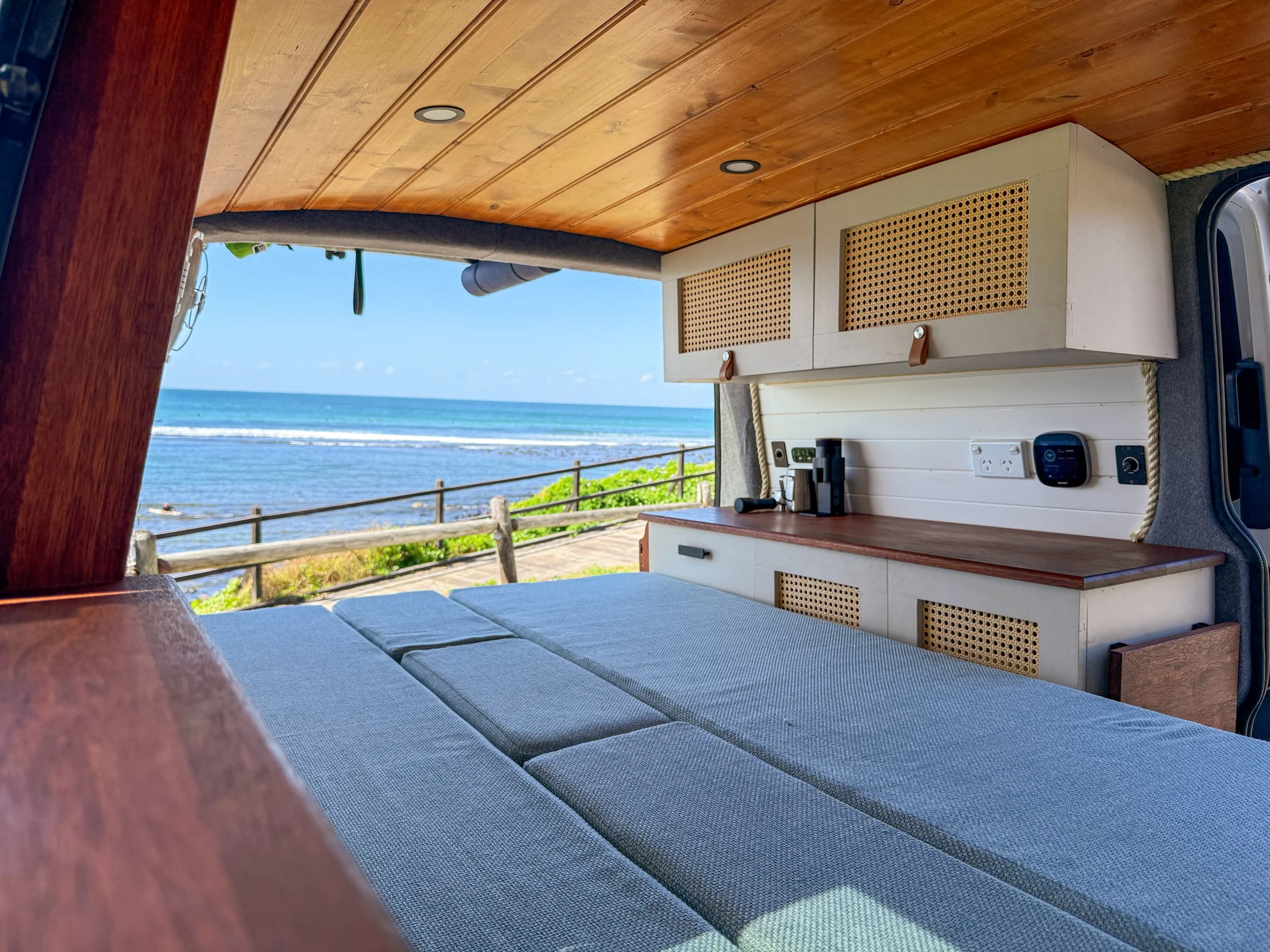 View from inside a camper van, looking out through an open side door at a beach and ocean with a clear blue sky, wooden railing, and green vegetation in the foreground.