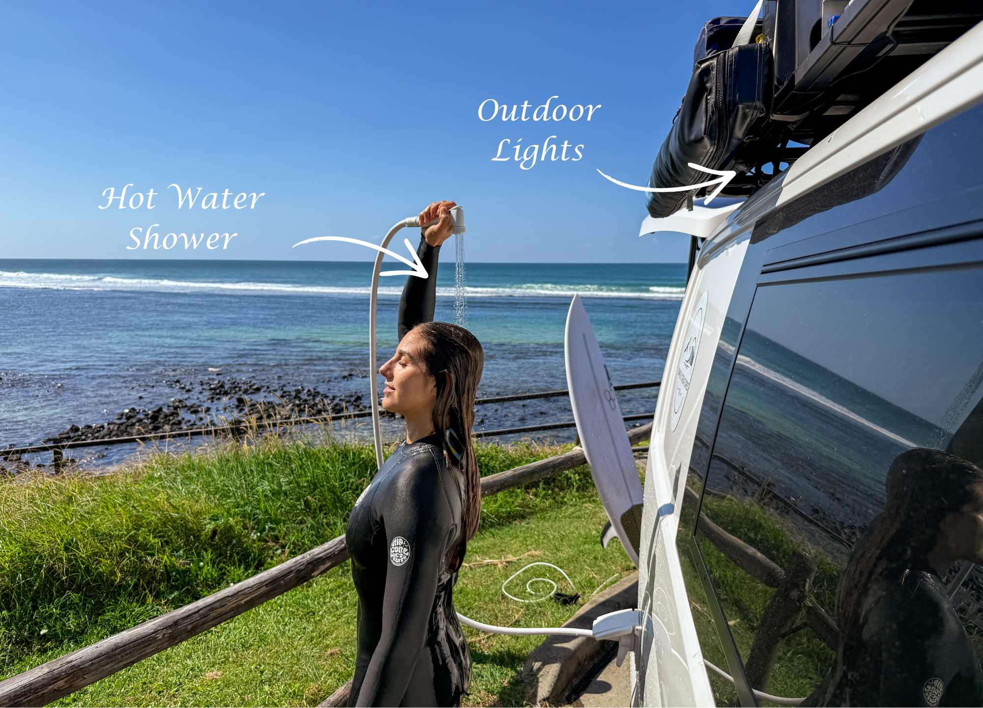 Woman in wetsuit standing on grass near ocean, holding showerhead with water, surfboard leaning against her, vehicle with outdoor lights and bag on roof rack in background.