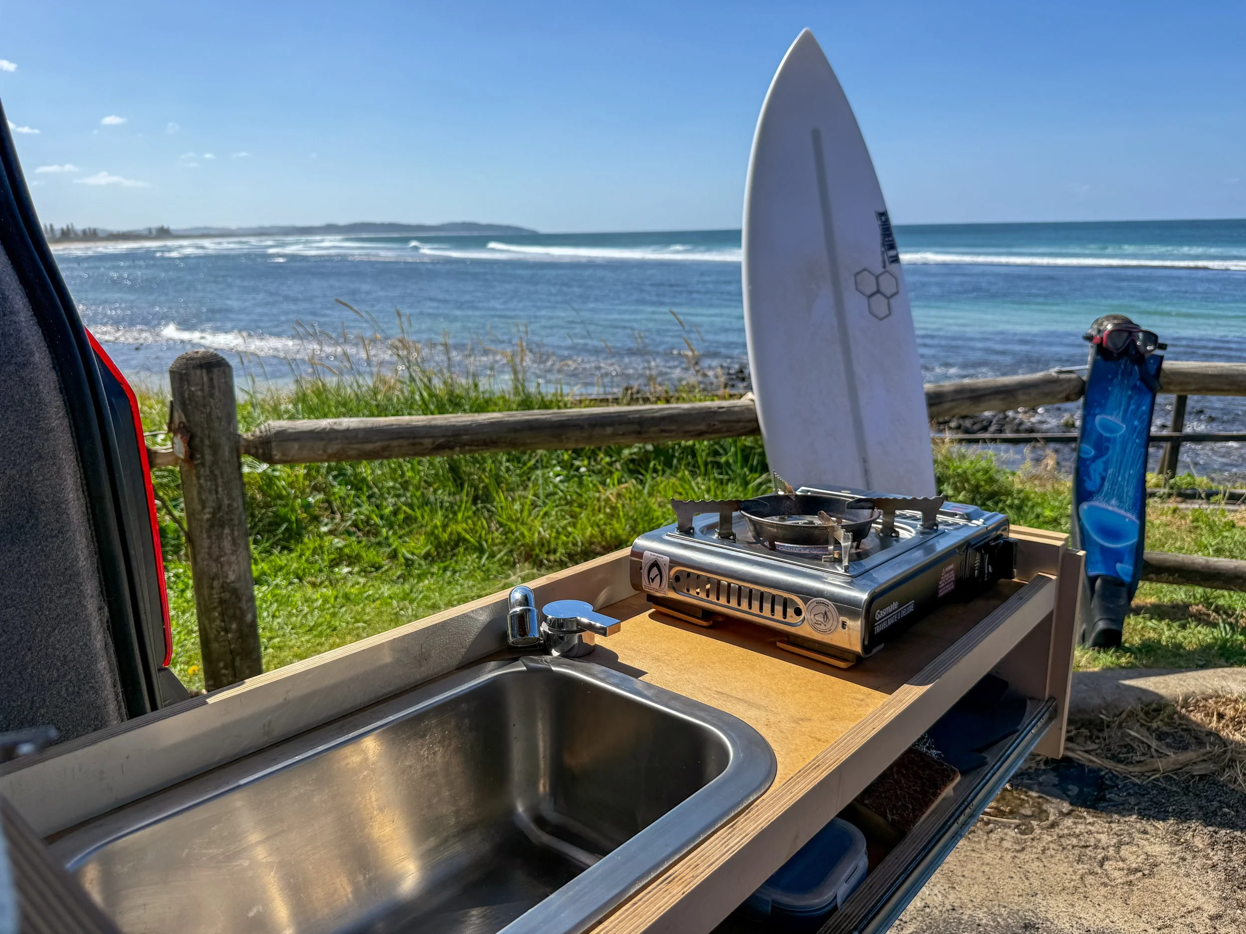 View of an ocean coastline with waves, taken from an outdoor camping kitchen setup, including a portable gas stove, a metal sink, and a surfboard standing upright near a wooden fence, with blue sky and scattered clouds in the background.