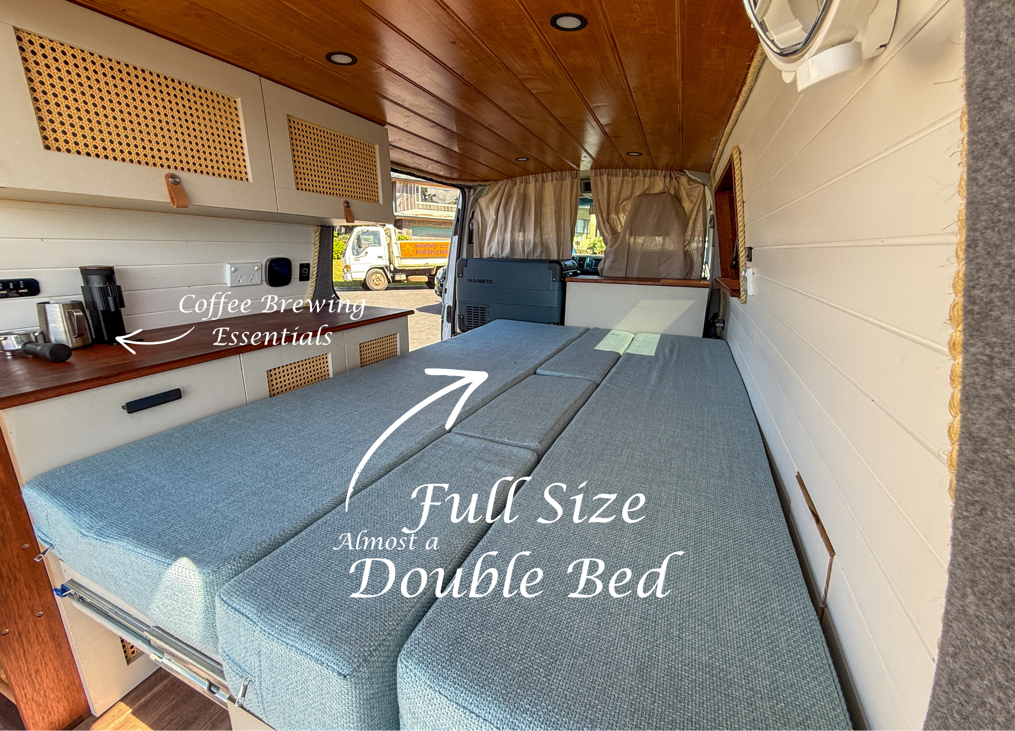 Interior of a camper van with a fold-out bed, a kitchenette area with coffee brewing essentials, and window curtains. The space is cozy with wood paneling and white walls.