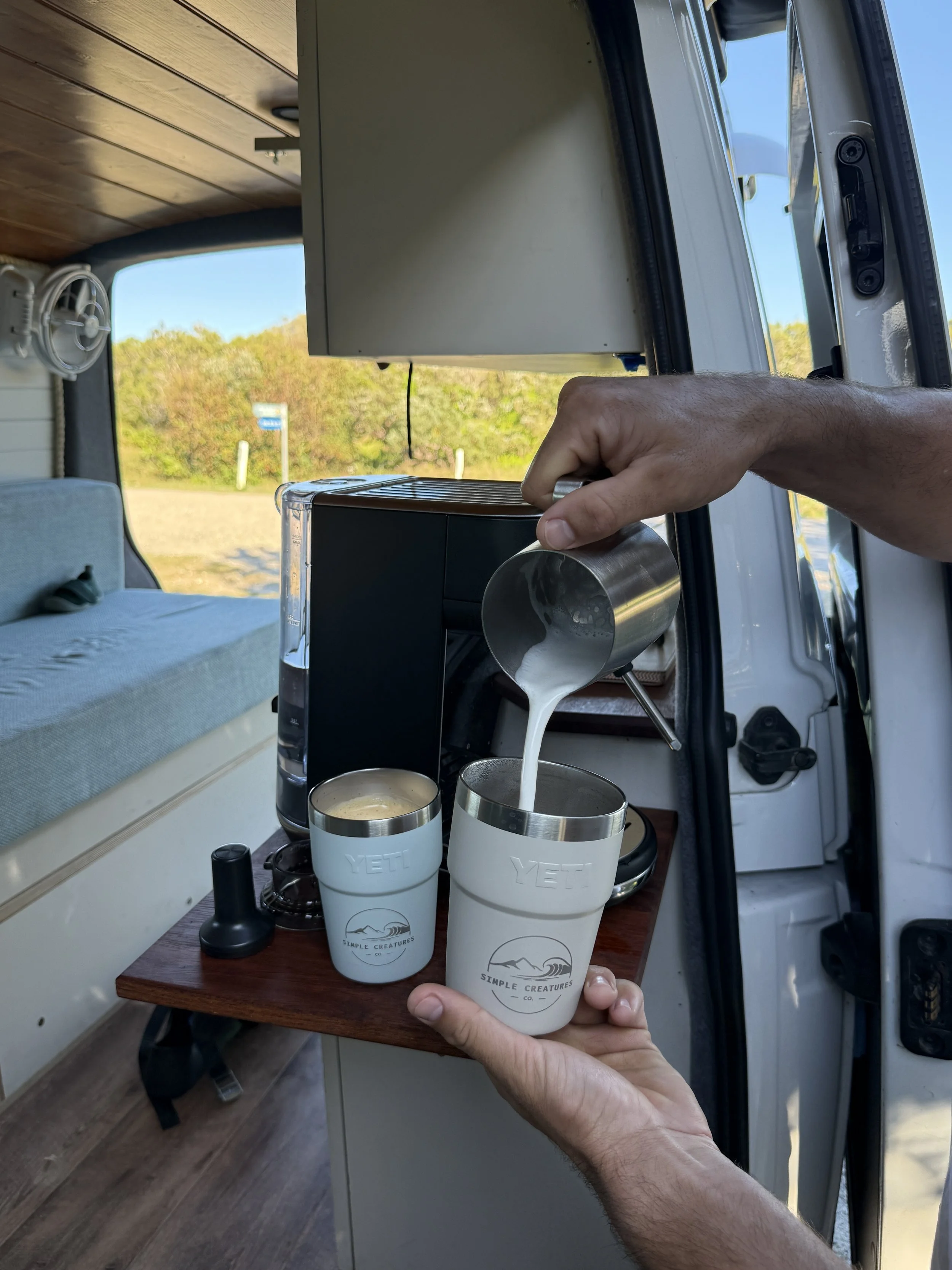 A person is holding two white YETI cups with a mountain logo and 'Simple Creatures Co.' printed on them, while pouring a creamy beverage into one of the cups inside a vehicle with a scenic outdoor view through the open door.