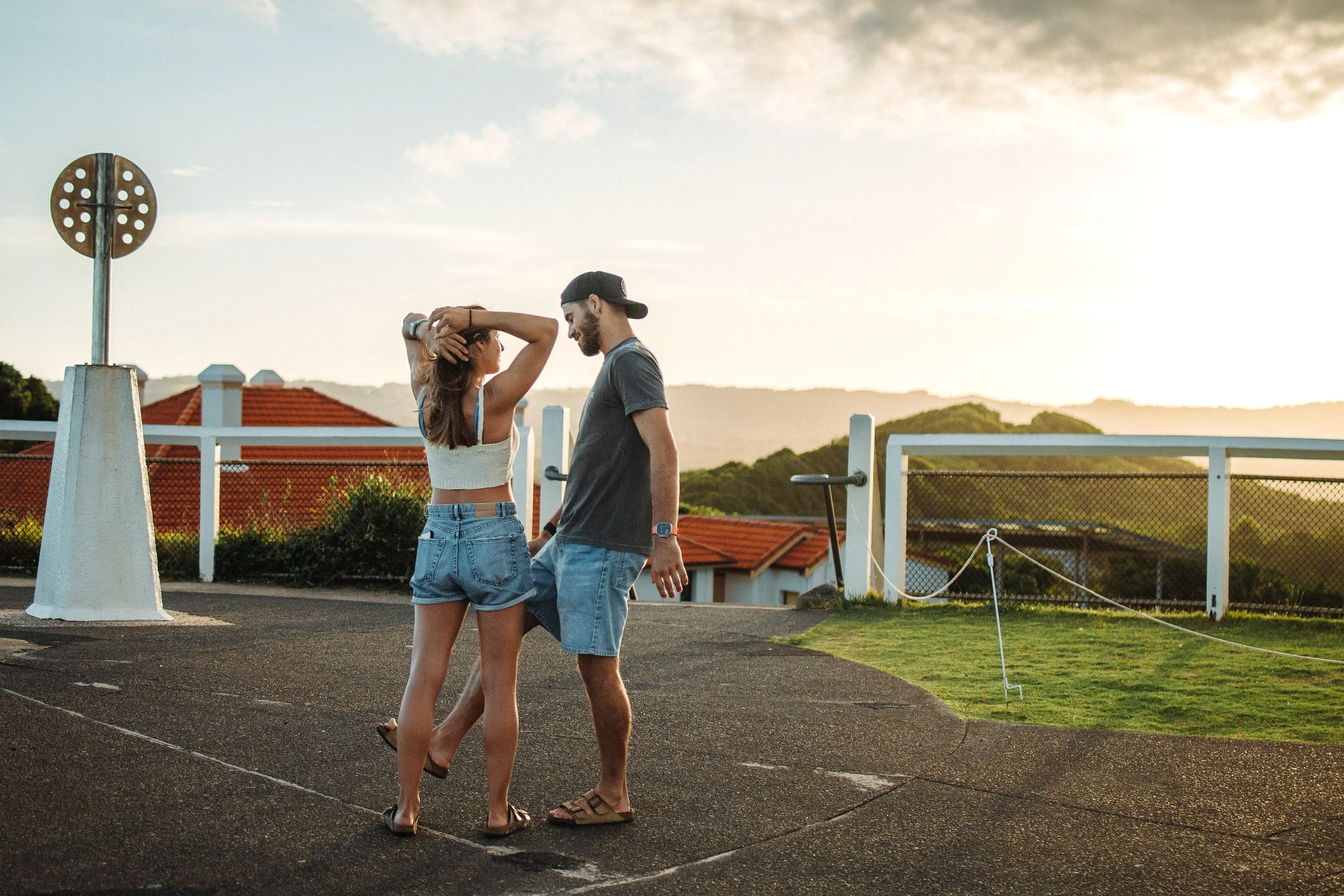 A young man and woman are standing close together outdoors during sunset, smiling and looking at each other. The woman has her hand on her head, and they appear to be enjoying a moment of intimacy. In the background, there are houses with red rooftops and hills.