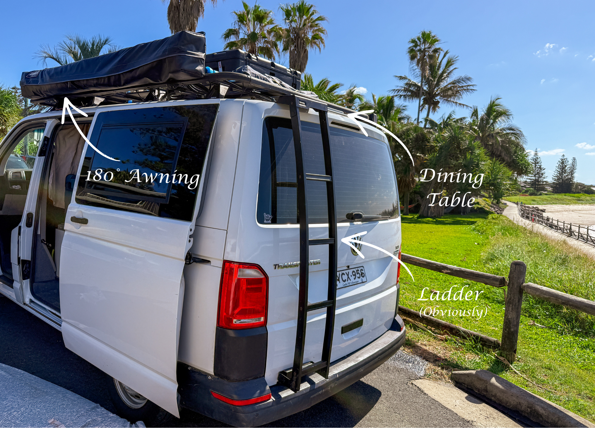 White van parked near a beach with palm trees in the background, featuring a mounted ladder on the back, an awning extended on the side, and a roof rack with a cargo box and a rooftop tent.