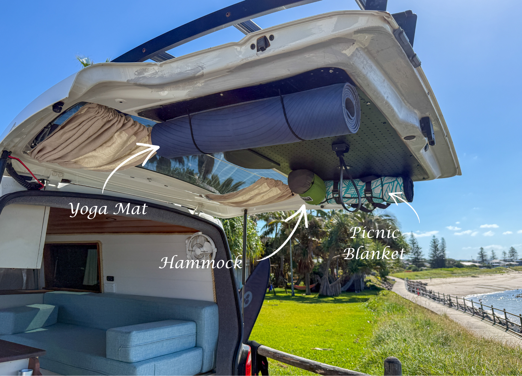 View of a van with its roof open, showing a hammock, yoga mat, picnic blanket, and attached accessories, parked near a grassy park with trees and a pathway beside water under a blue sky.