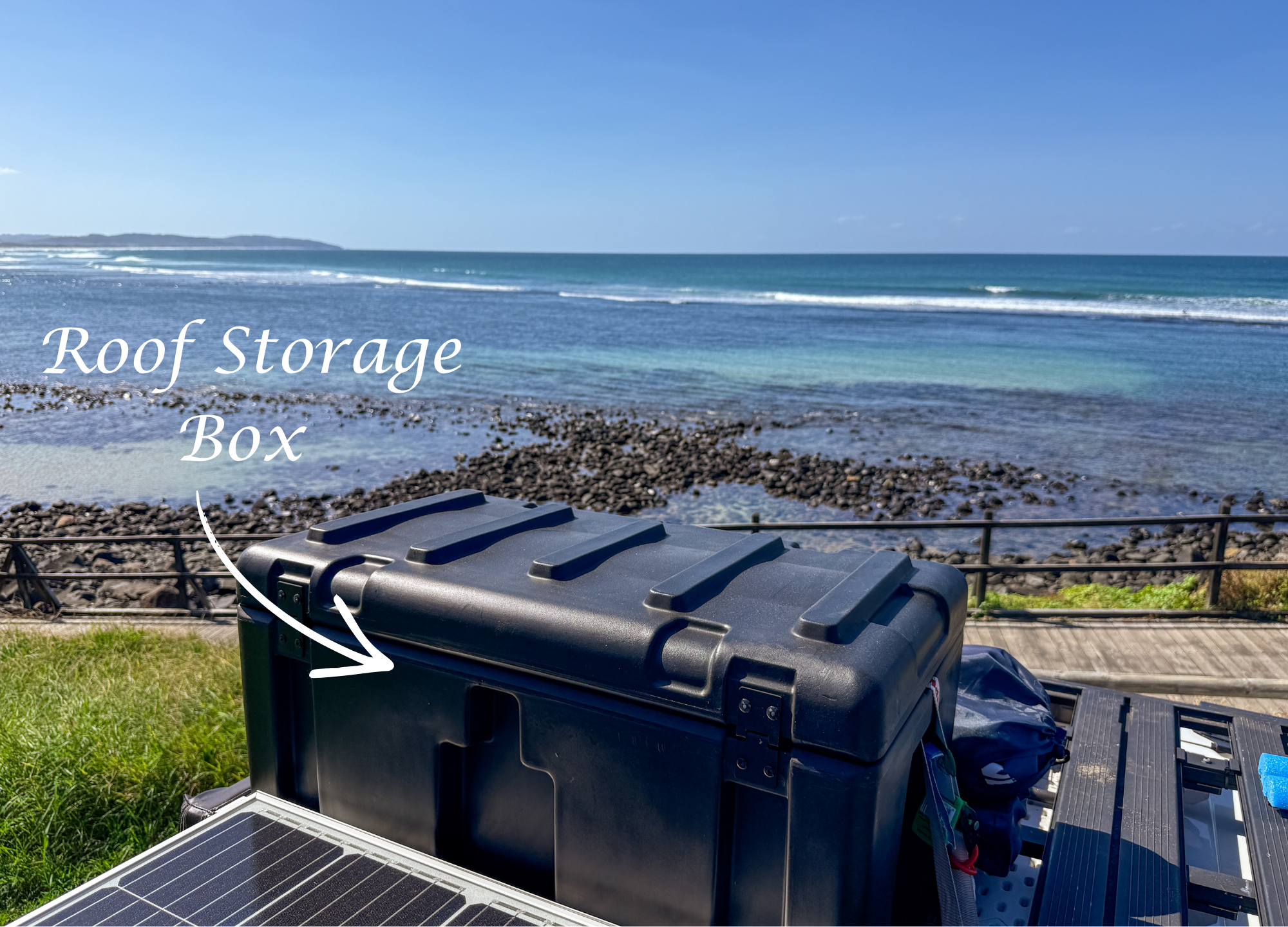A black portable solar generator with solar panels, a scenic ocean view with rocks, waves, and a blue sky, and a trash bin in the foreground.