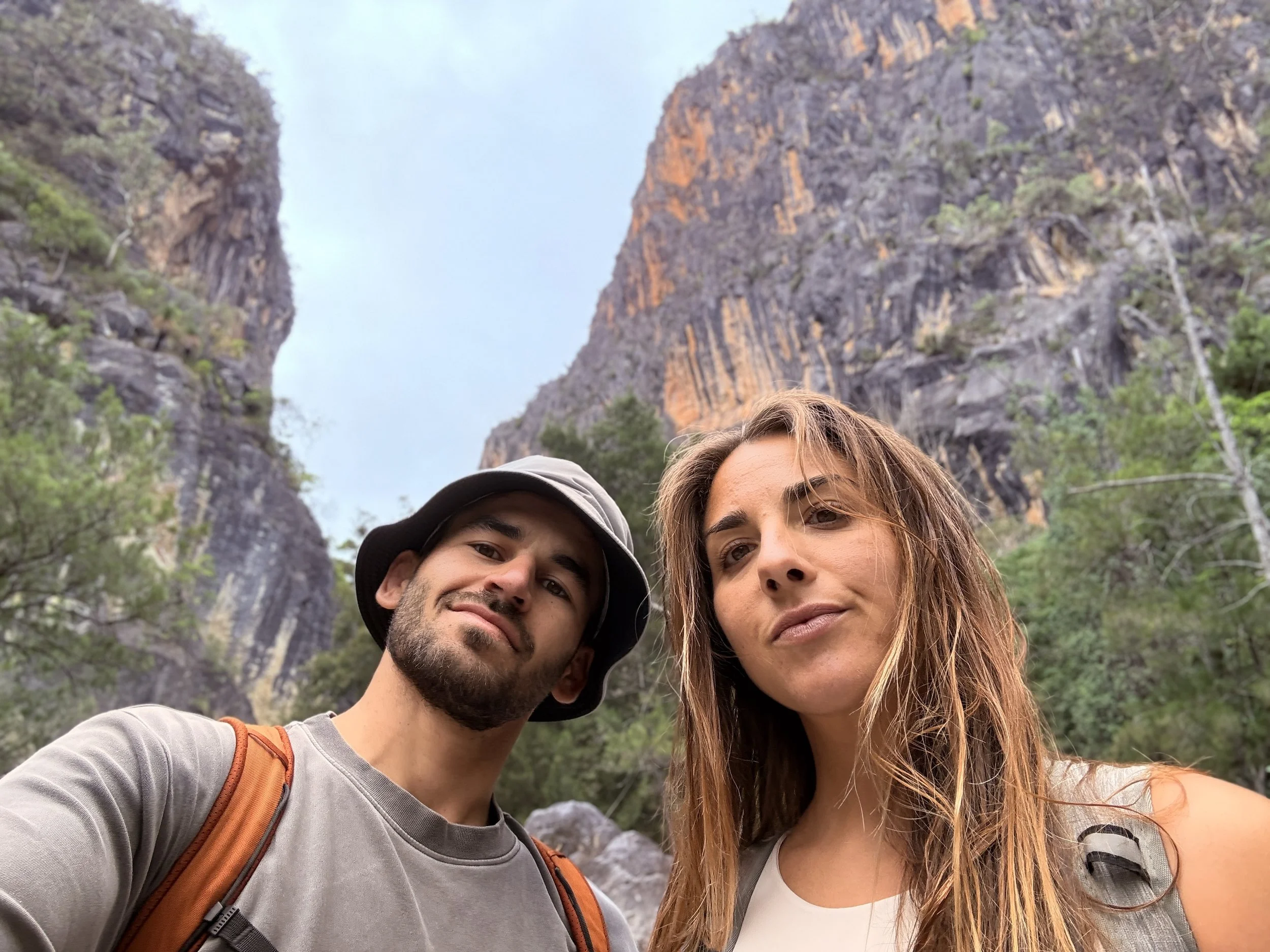 Two individuals taking a selfie outdoors in a scenic mountainous area with rocky cliffs and sparse greenery in the background.