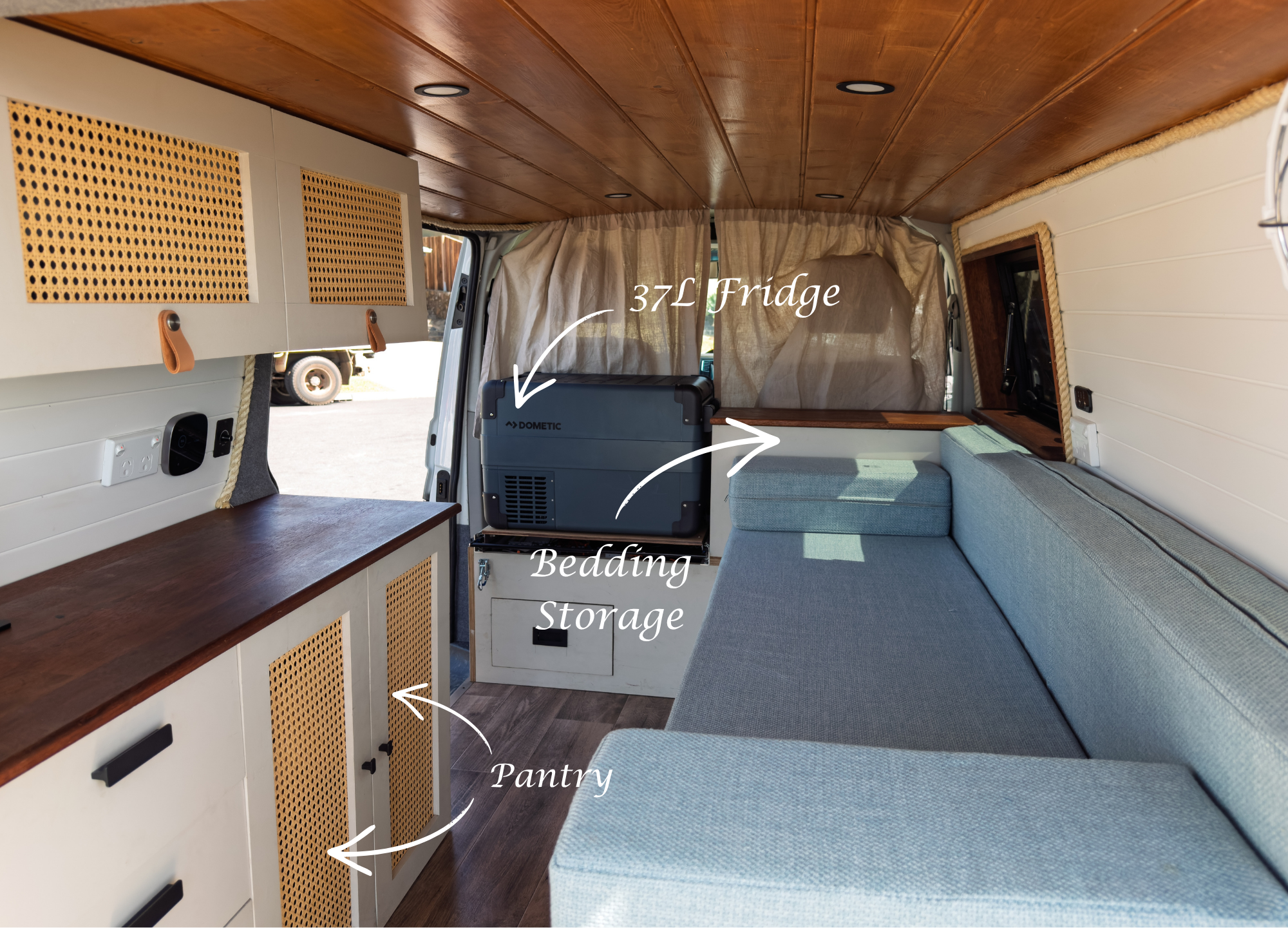 Interior of a camper van showing cabinetry, a pantry, a fridge, and seating with curtains and wooden ceiling.