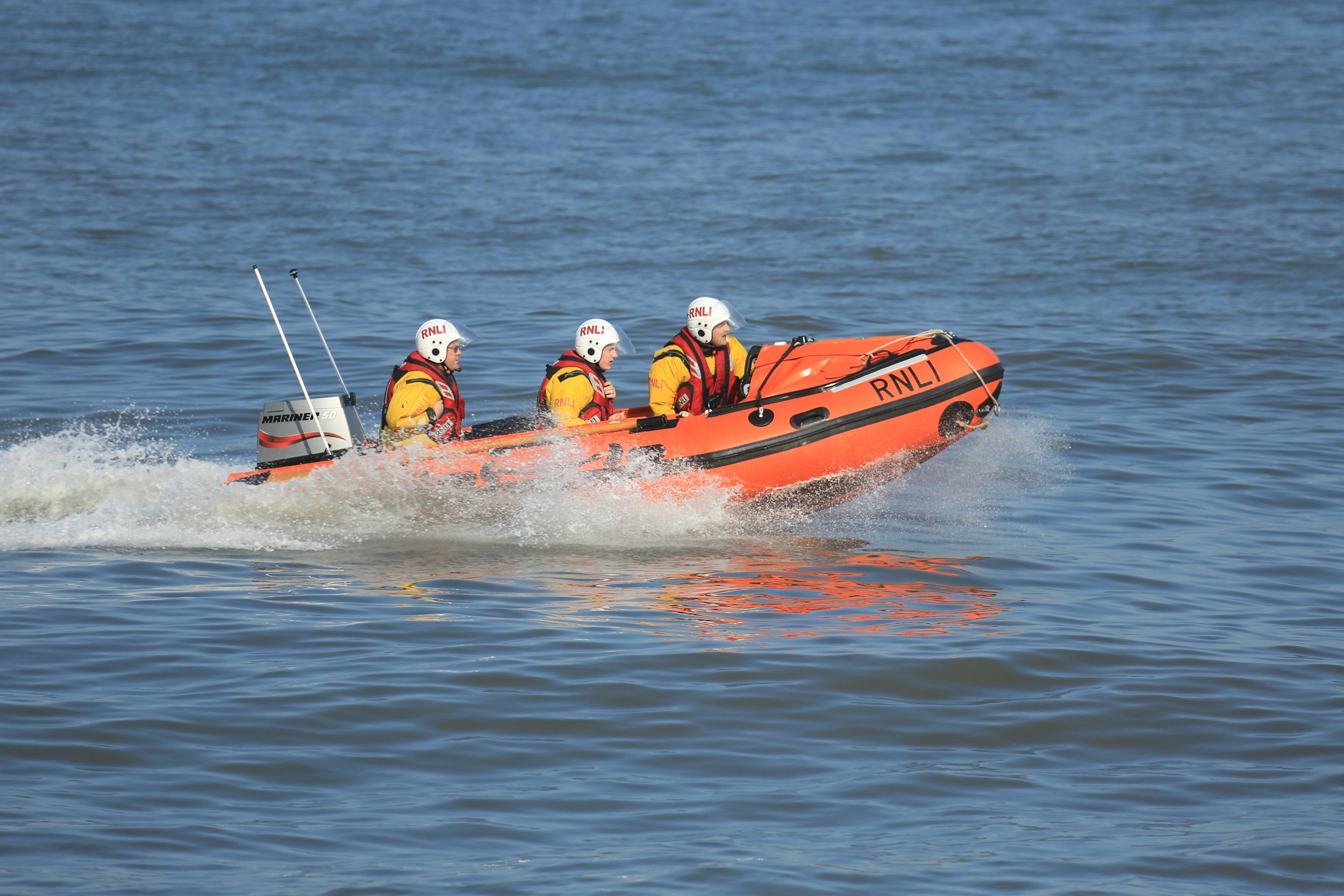 Three rescue team members in a small orange boat speeding across the water, wearing helmets and yellow uniforms with RNLi markings.