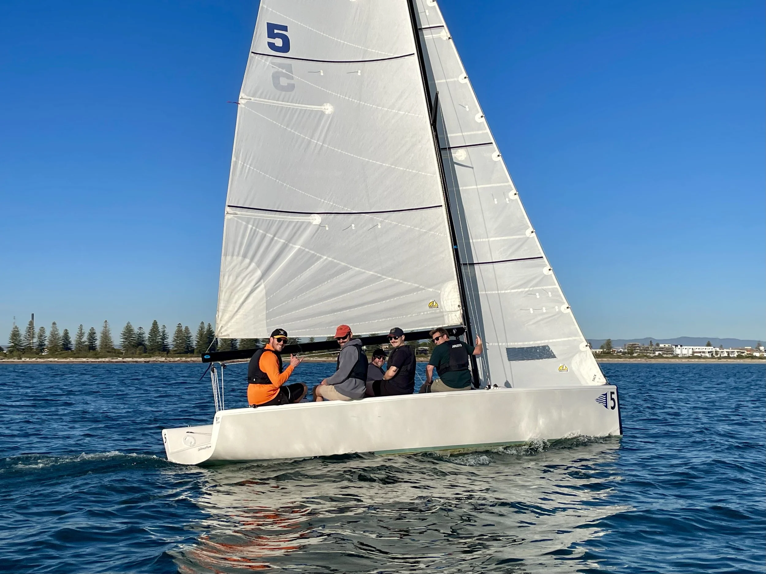 A sailboat with four people on board sailing on the water under a clear blue sky.