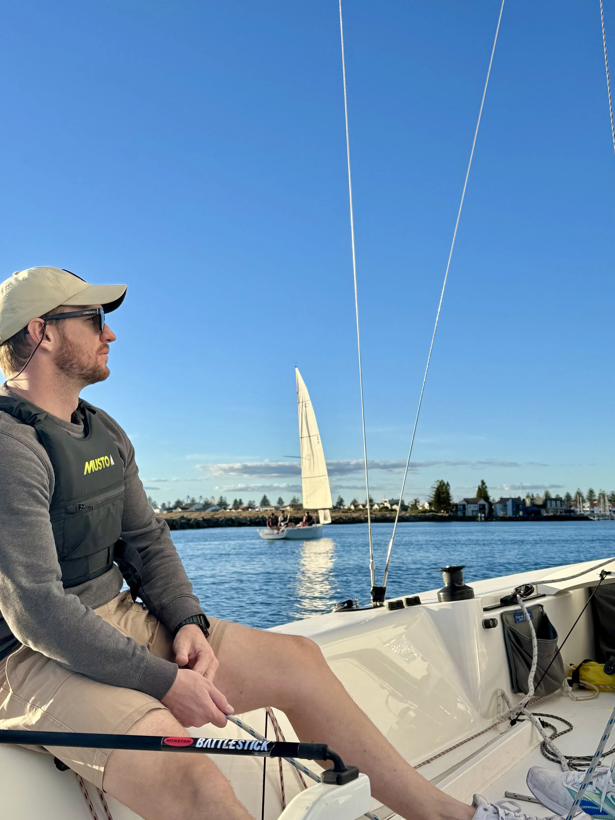 A man sitting on a sailboat, wearing sunglasses, a beige cap, a black Life vest with yellow text, and shorts, looking at the water with a sailboat in the background on a clear, sunny day.