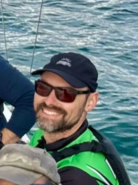 A man smiling on a boat, wearing sunglasses, a black cap, and a green life jacket, with water in the background.