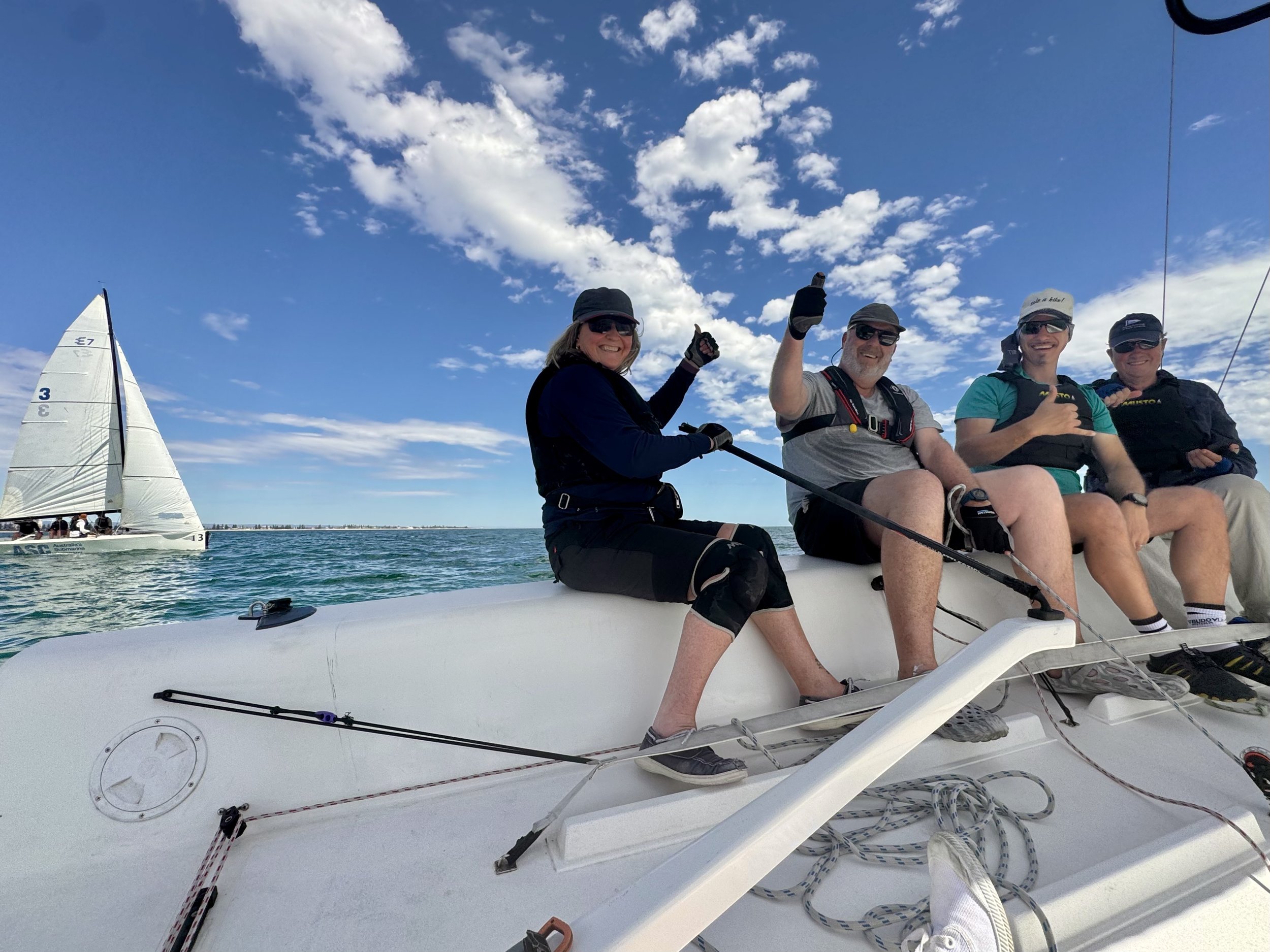 Four people sitting and smiling on a sailboat under a blue sky with white clouds, with a yacht sailing in the background.