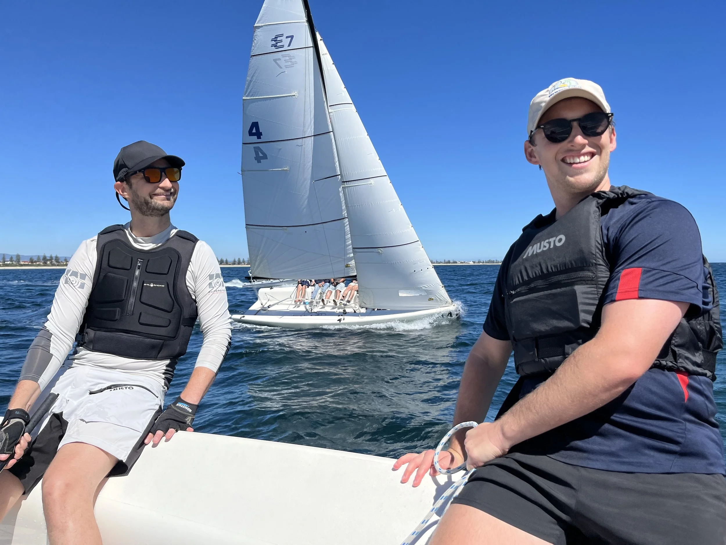 Two smiling men with sunglasses and life jackets on a sailboat, sailing on a clear day with a distant shoreline in the background.