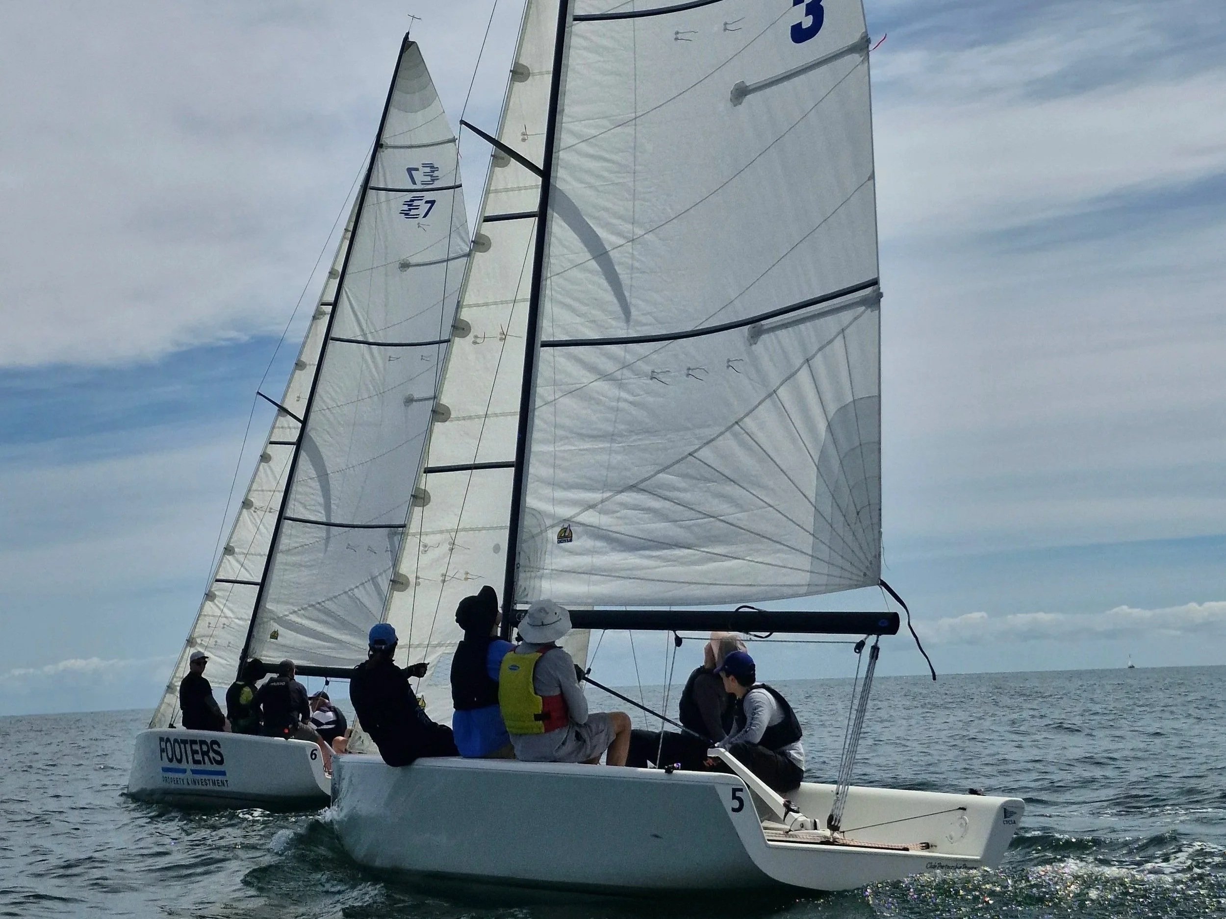 A group of people sailing on a catamaran with white sails on a calm ocean with a partly cloudy sky in the background.