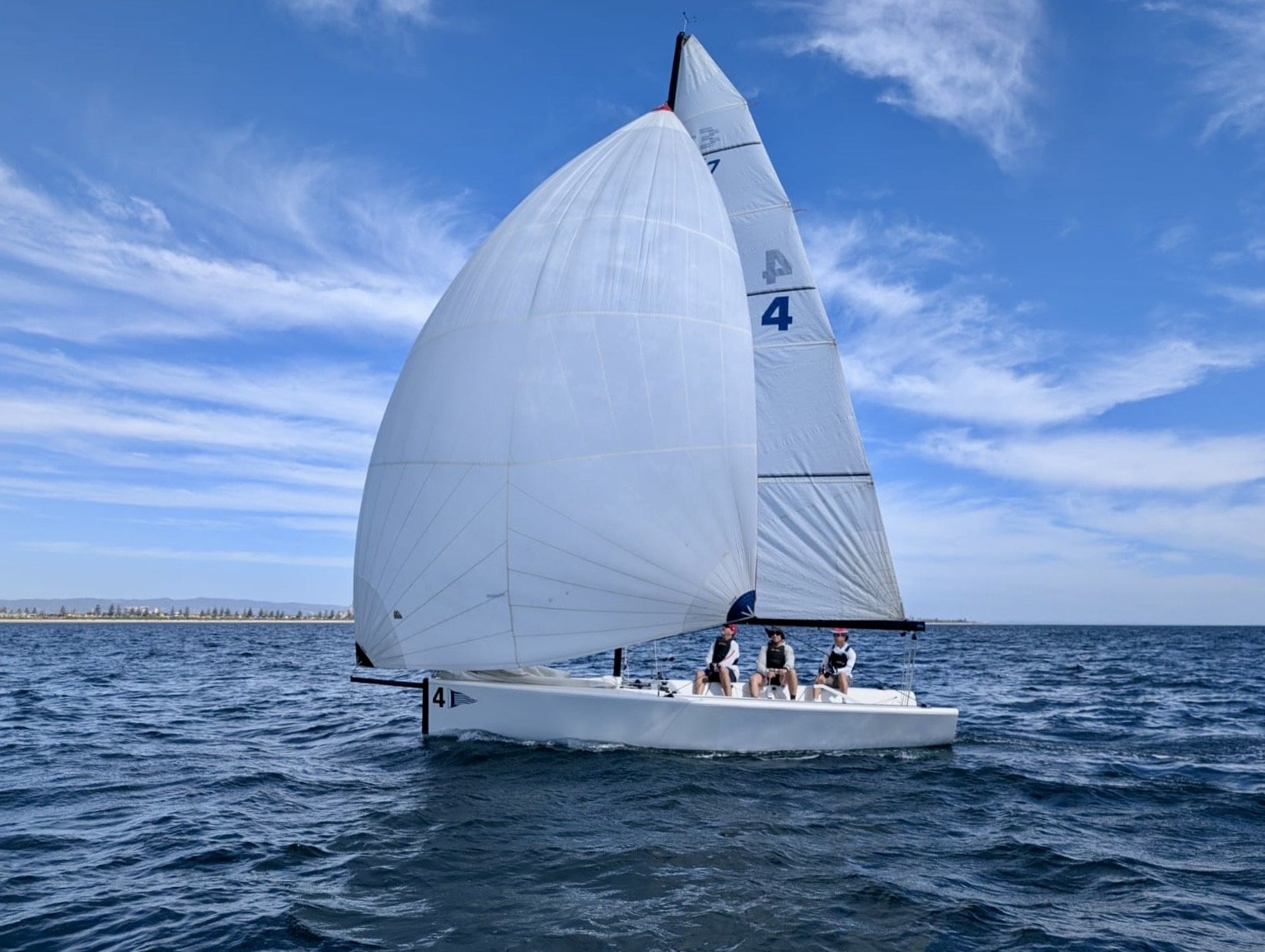 A sailboat with a large white spinnaker sail and three crew members sailing on the water under a partly cloudy sky.