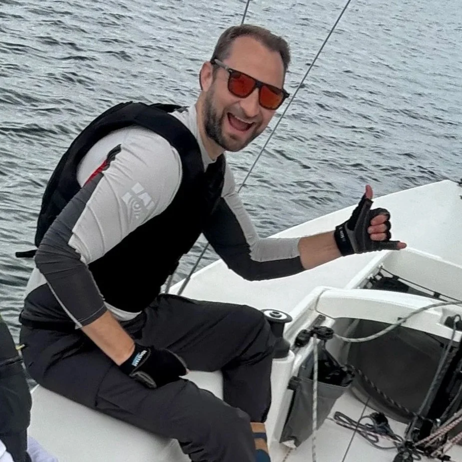 A man smiling and wearing sunglasses, gloves, and a backpack, sitting on a boat with water in the background, giving a shaka sign.