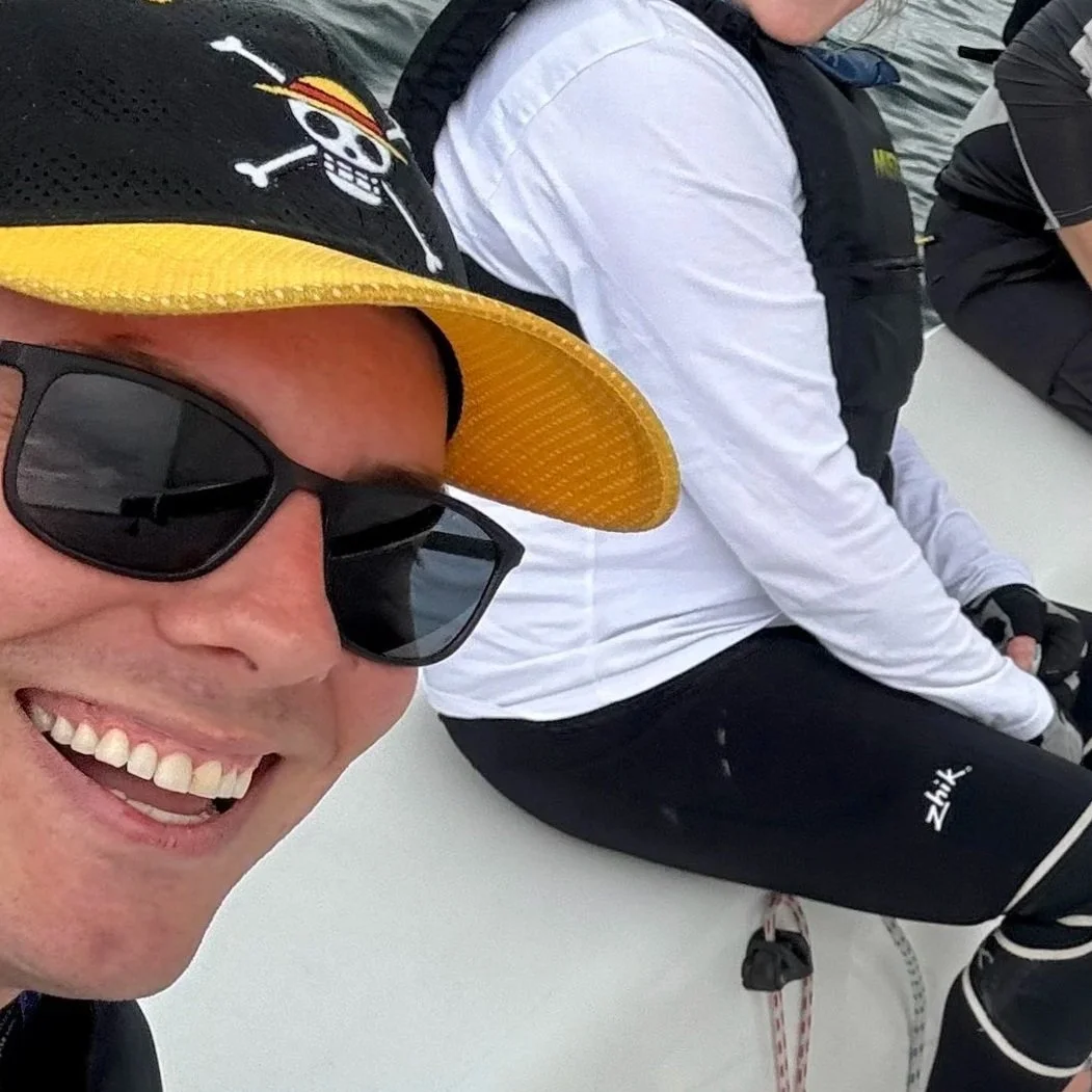 Close-up of a smiling man wearing sunglasses and a black cap with a skull and crossbones design, sitting on a boat with others.