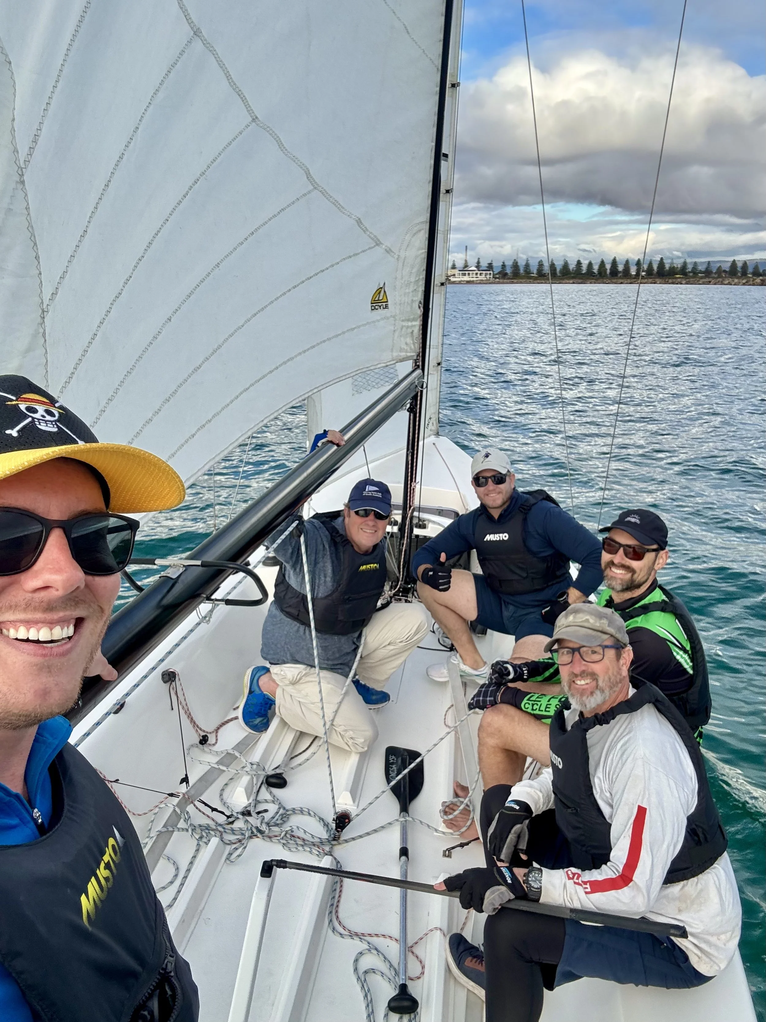 Group of five crew sailing on a boat, smiling, wearing life jackets and sunglasses, with a body of water and cloudy sky in the background.