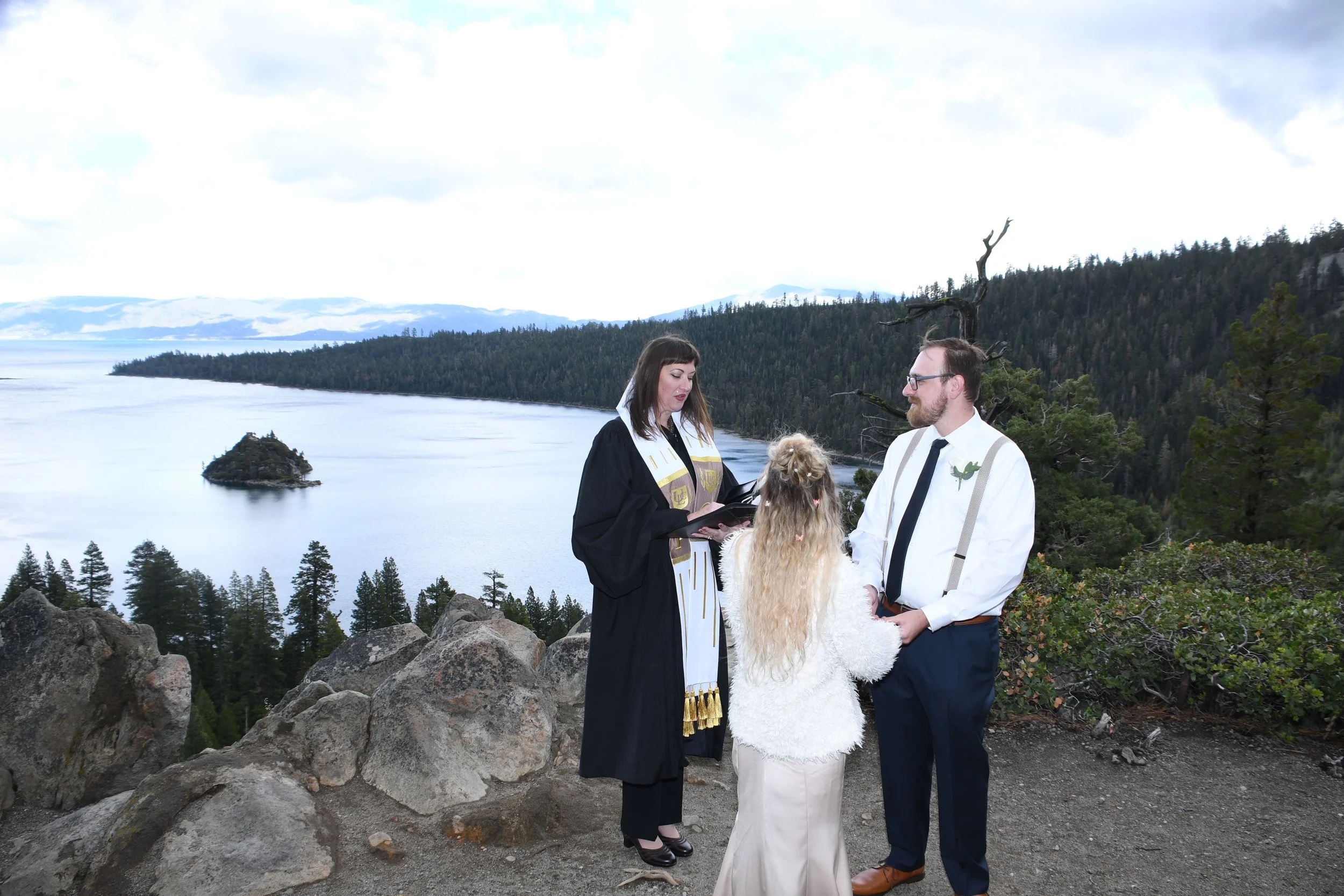 A wedding ceremony taking place outdoors on a rocky ledge overlooking a lake and forested mountains. A woman in a wedding dress with long blonde hair, a man in a white shirt with suspenders and tie, and a woman in a black coat are participating in th