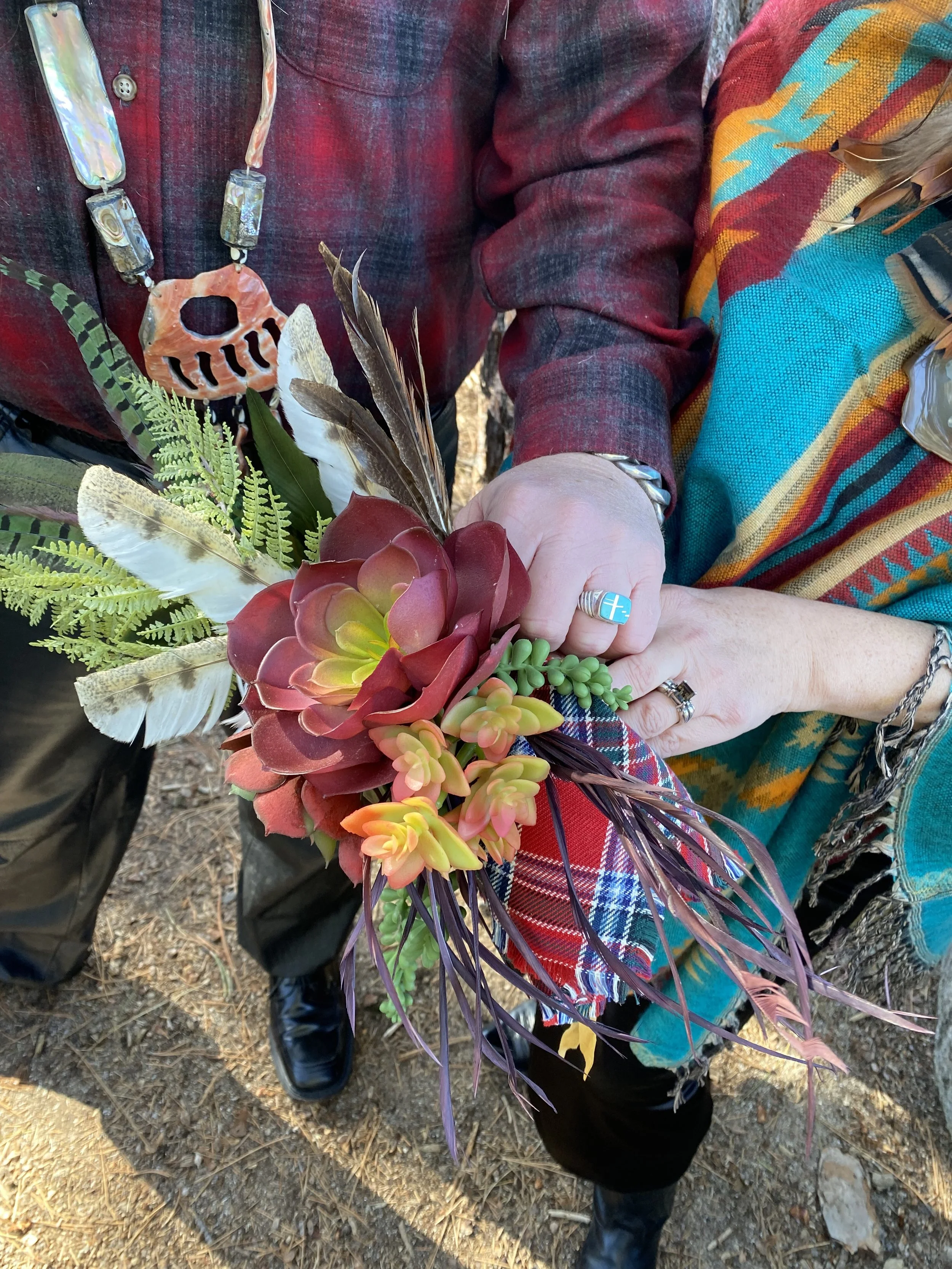 Bride holding a bouquet of artificial succulents and feathers, wearing a plaid shirt and a colorful scarf, with a beaded necklace and showing wedding rings.