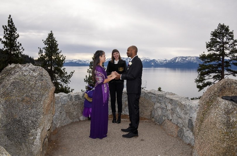 A wedding ceremony taking place outdoors with a couple holding hands and an officiant standing behind them, overlooking a lake and snow-capped mountains.