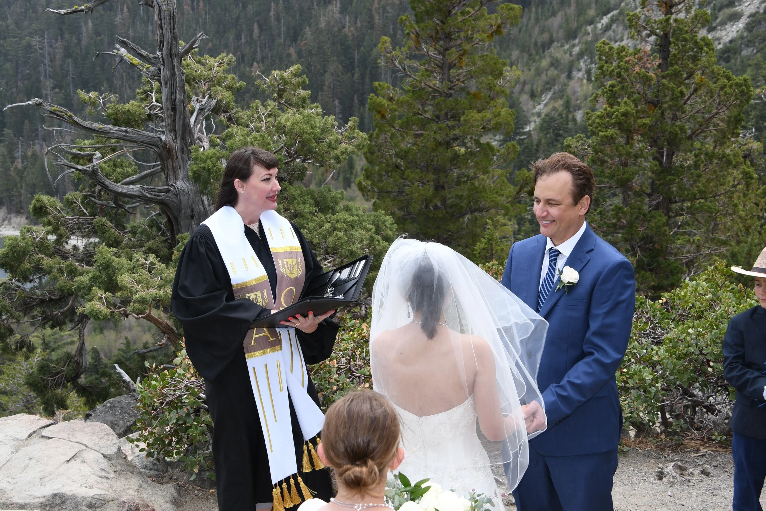 A couple gets married outdoors with a mountain and trees in the background, officiant reading from a book, bride in white with veil, groom in blue suit, and a young girl with floral hair accessory.